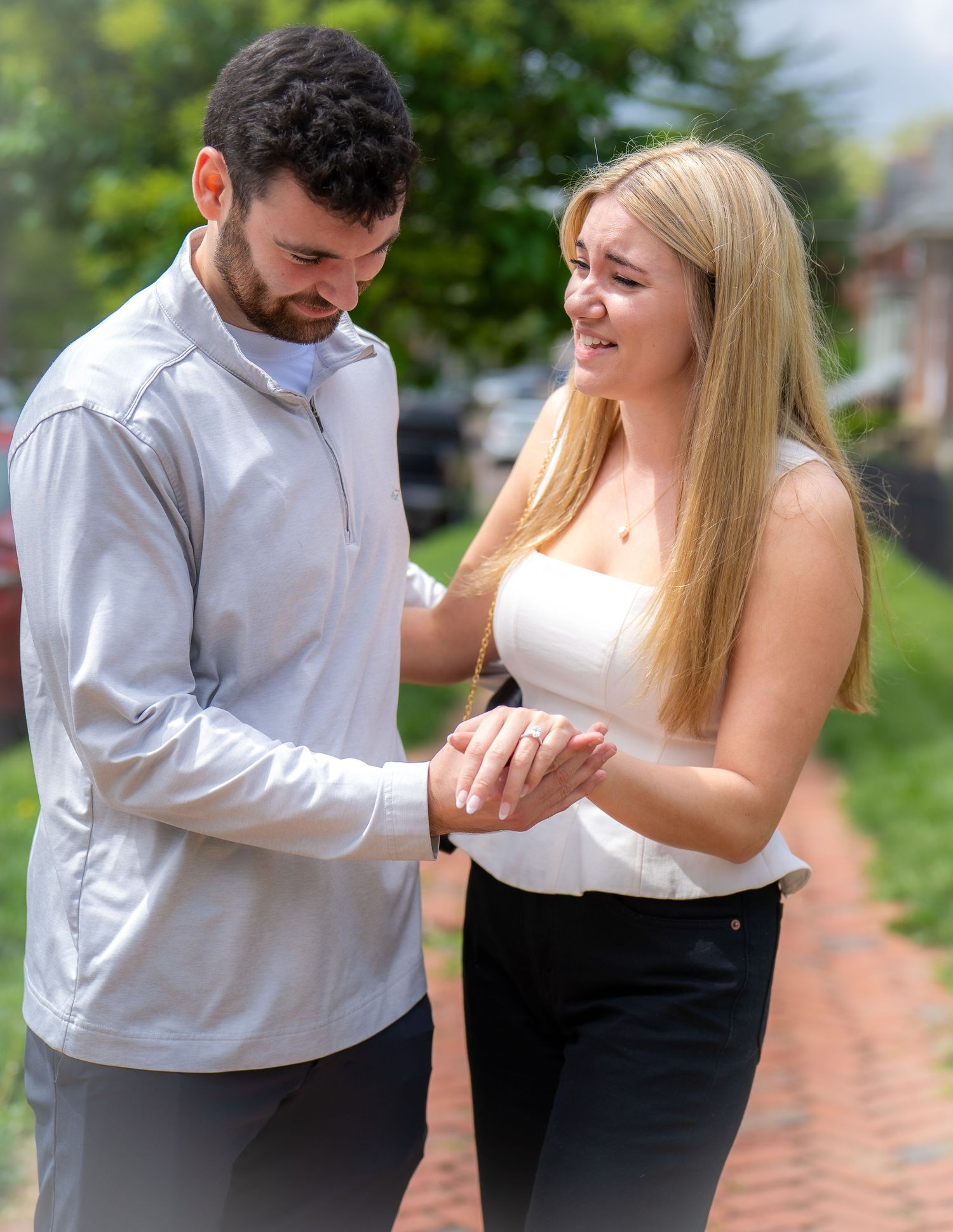 Joyful newly engaged couple embracing and smiling outdoors with green trees behind them.