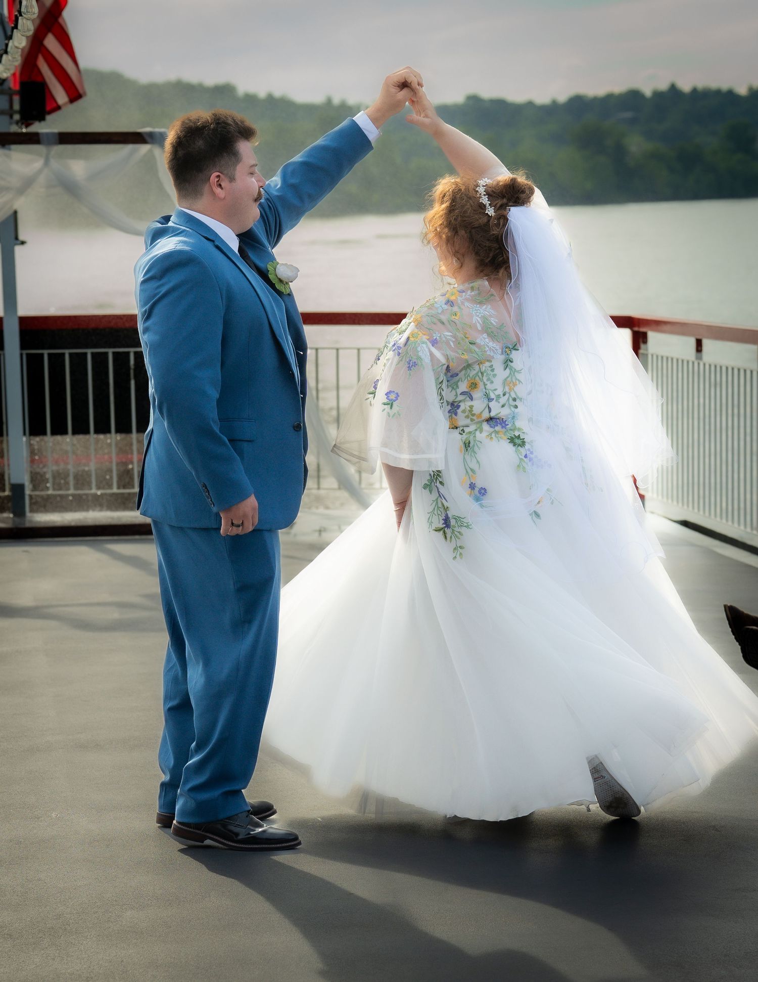Bride and groom dancing on boat deck, bride in floral white gown with veil, groom in blue suit, water background.