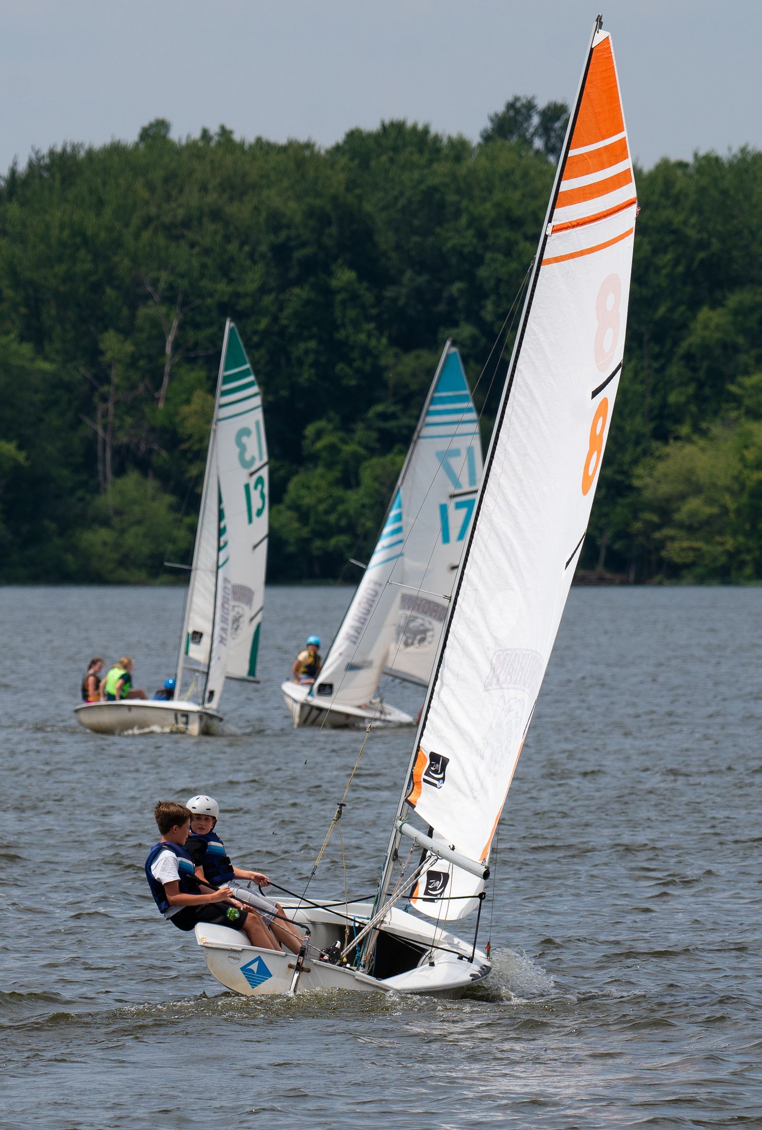 Youth sailors race small sailboats with colorful sails across a lake during sailing competition.