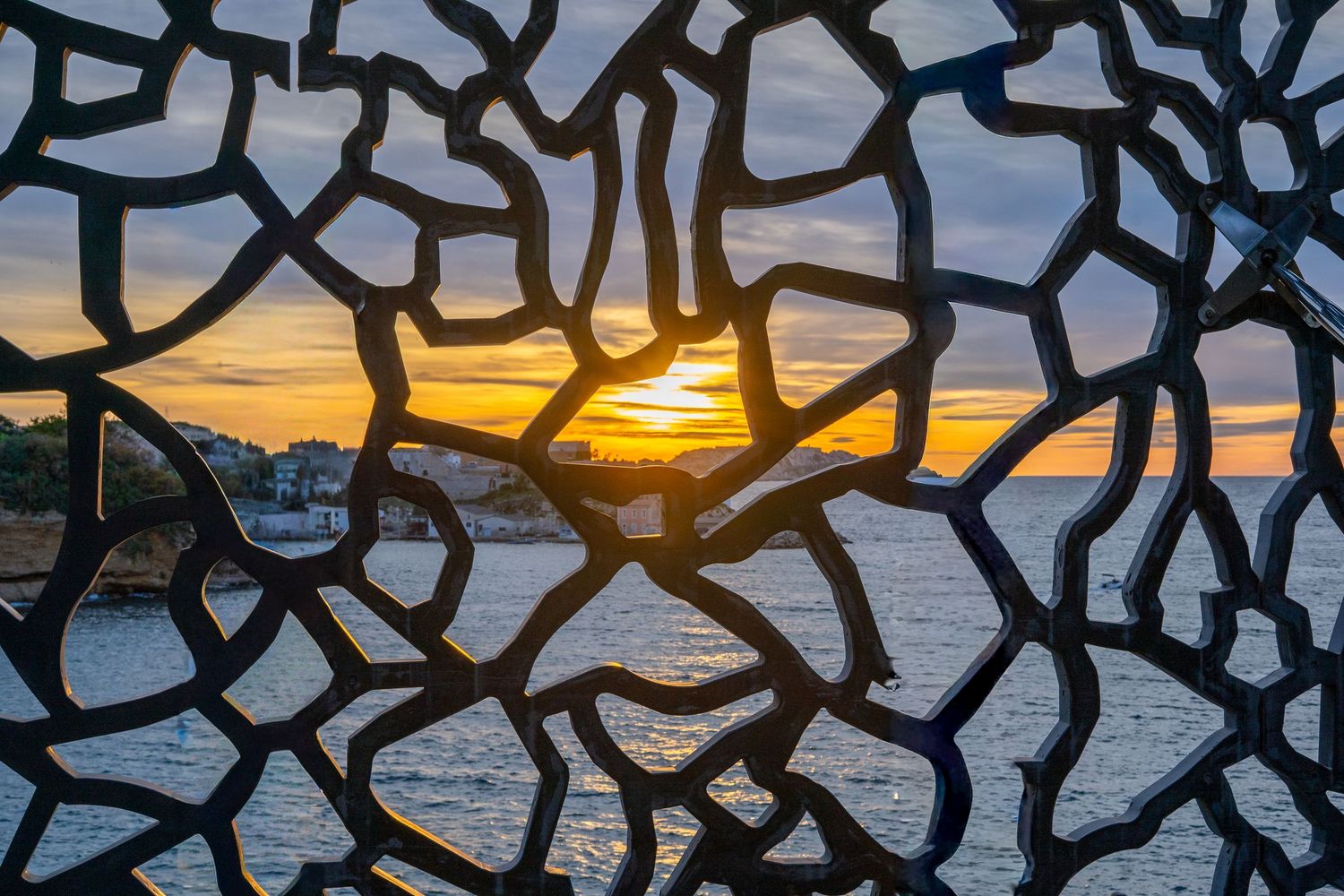 Sunset viewed through intricate metal lattice sculpture overlooking a calm ocean coastline at golden hour.