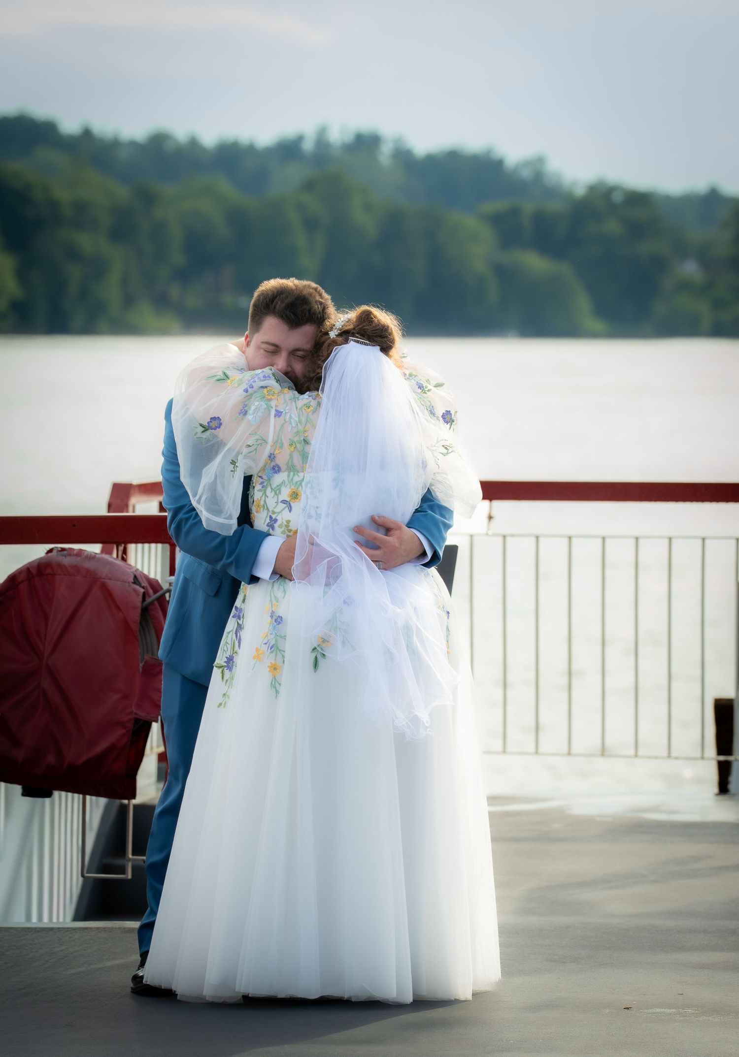 Newlywed couple embracing in wedding attire on a waterfront deck, bride in floral white gown with veil.