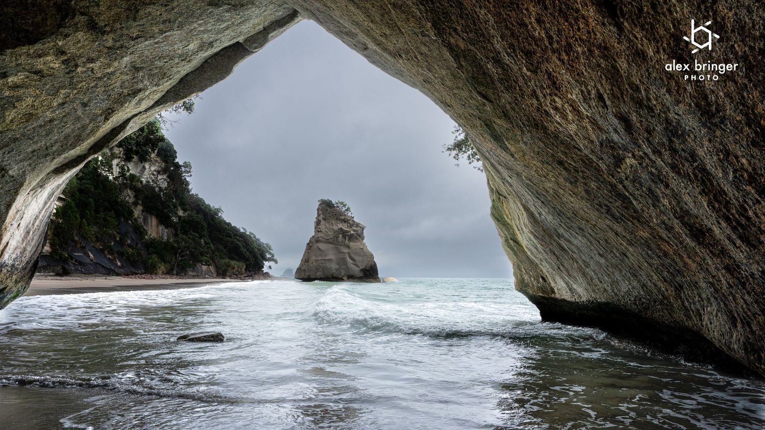 Natural rock arch frames a dramatic ocean view with a lone sea stack rising from the waves on a cloudy day.