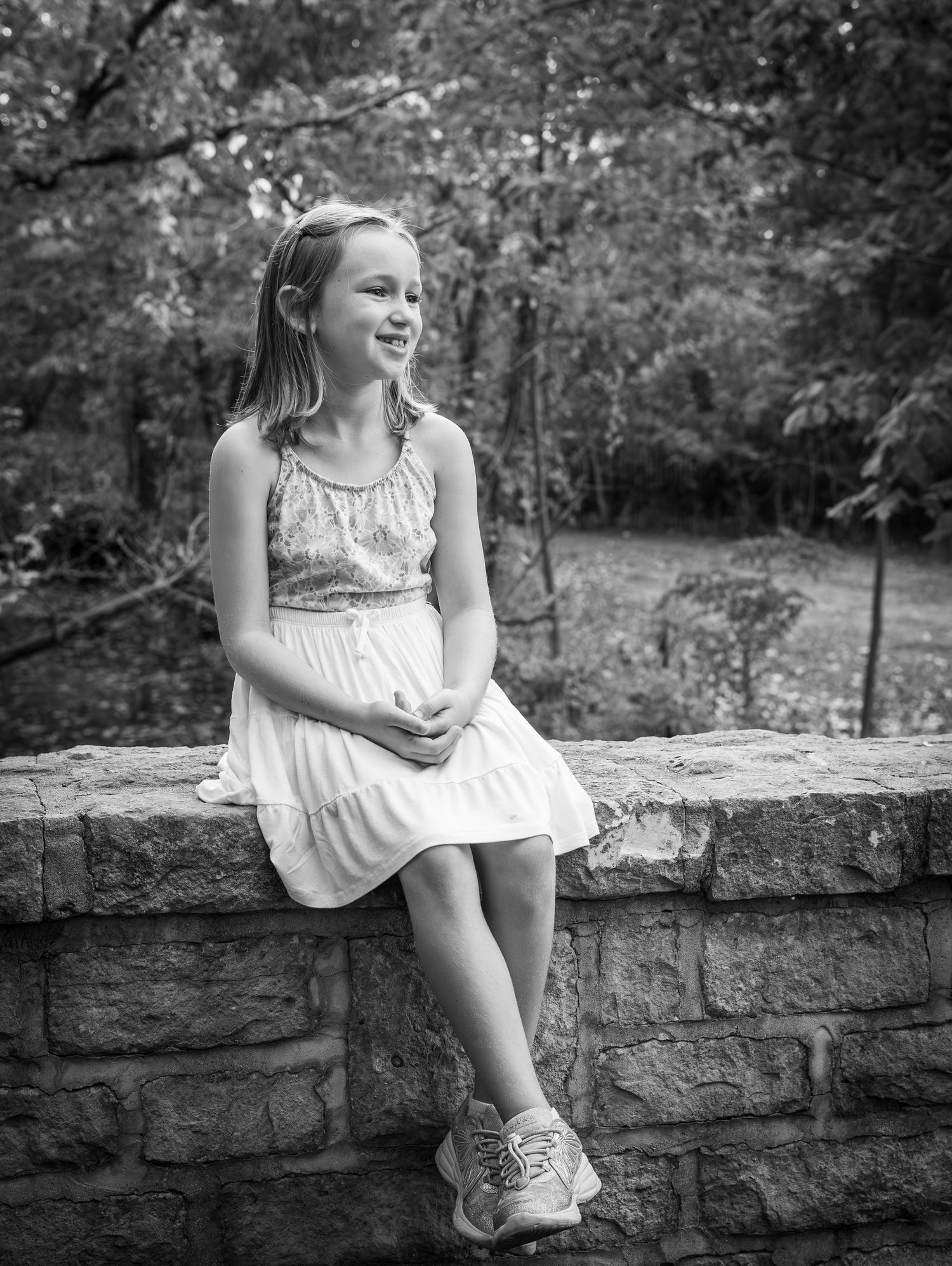 Smiling young girl in white dress sitting on a stone wall surrounded by trees in black and white photo.