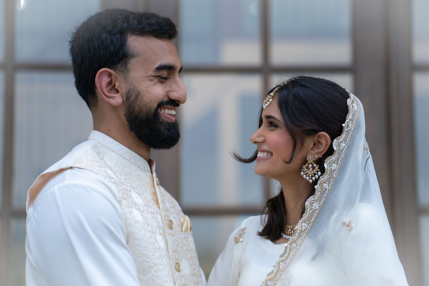 South Asian couple in white traditional wedding attire smiling at each other during nikah ceremony.