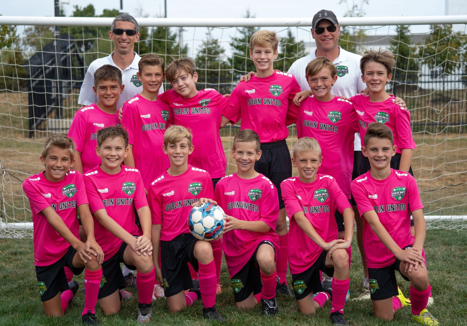 Youth soccer team in pink jerseys posing with coaches for a group photo on a soccer field.