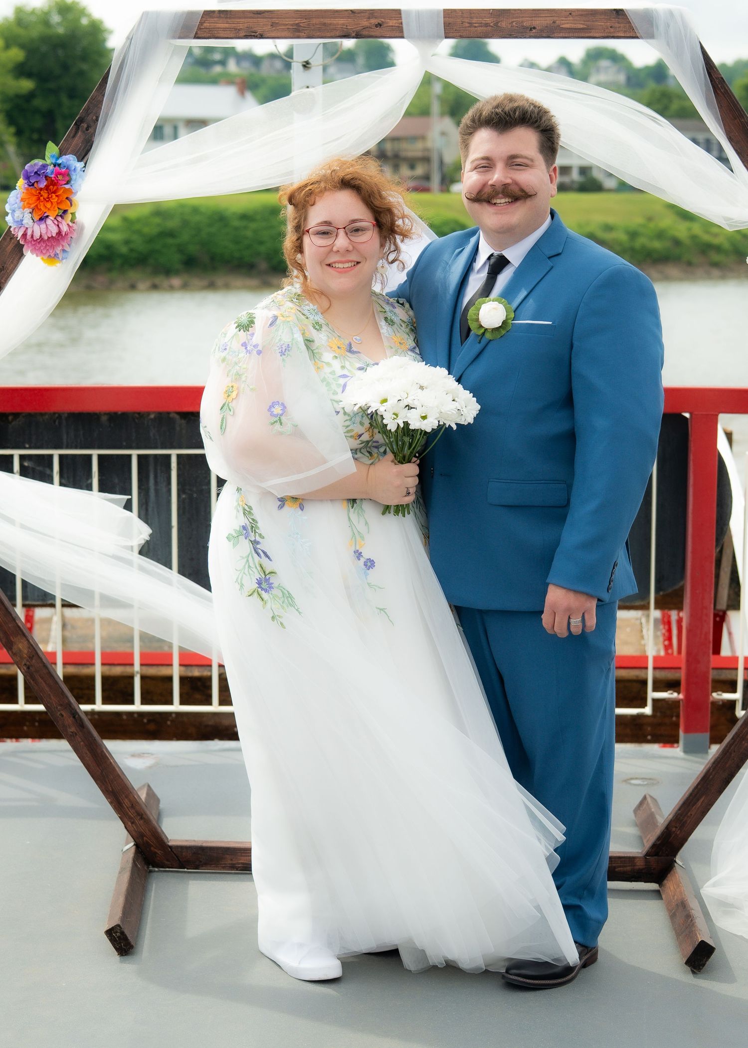 Bride and groom posing together outdoors on a waterfront deck, bride holding white flowers, groom in blue suit.