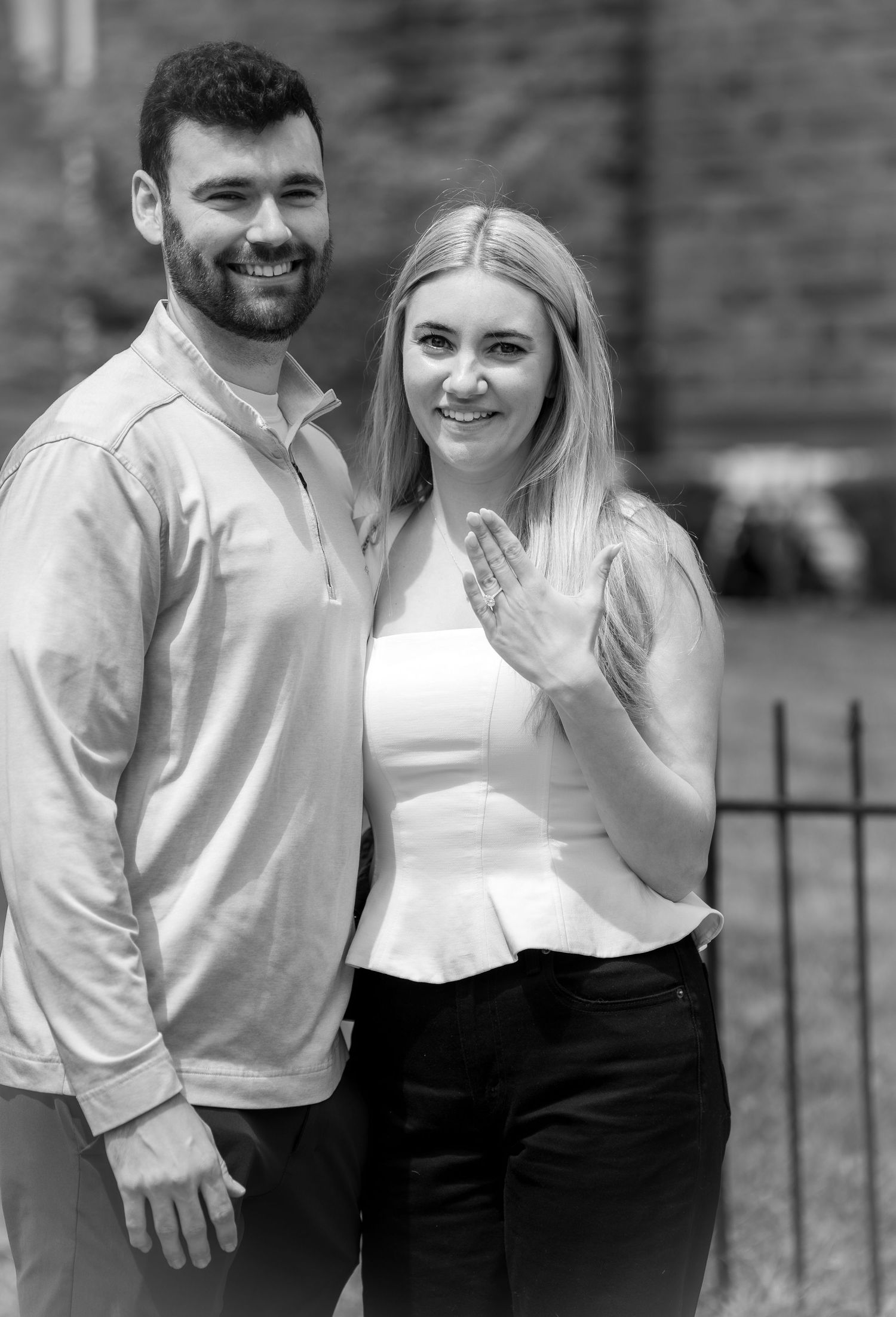 Black and white photo of a smiling couple posing outdoors, woman showing off engagement ring on her hand.