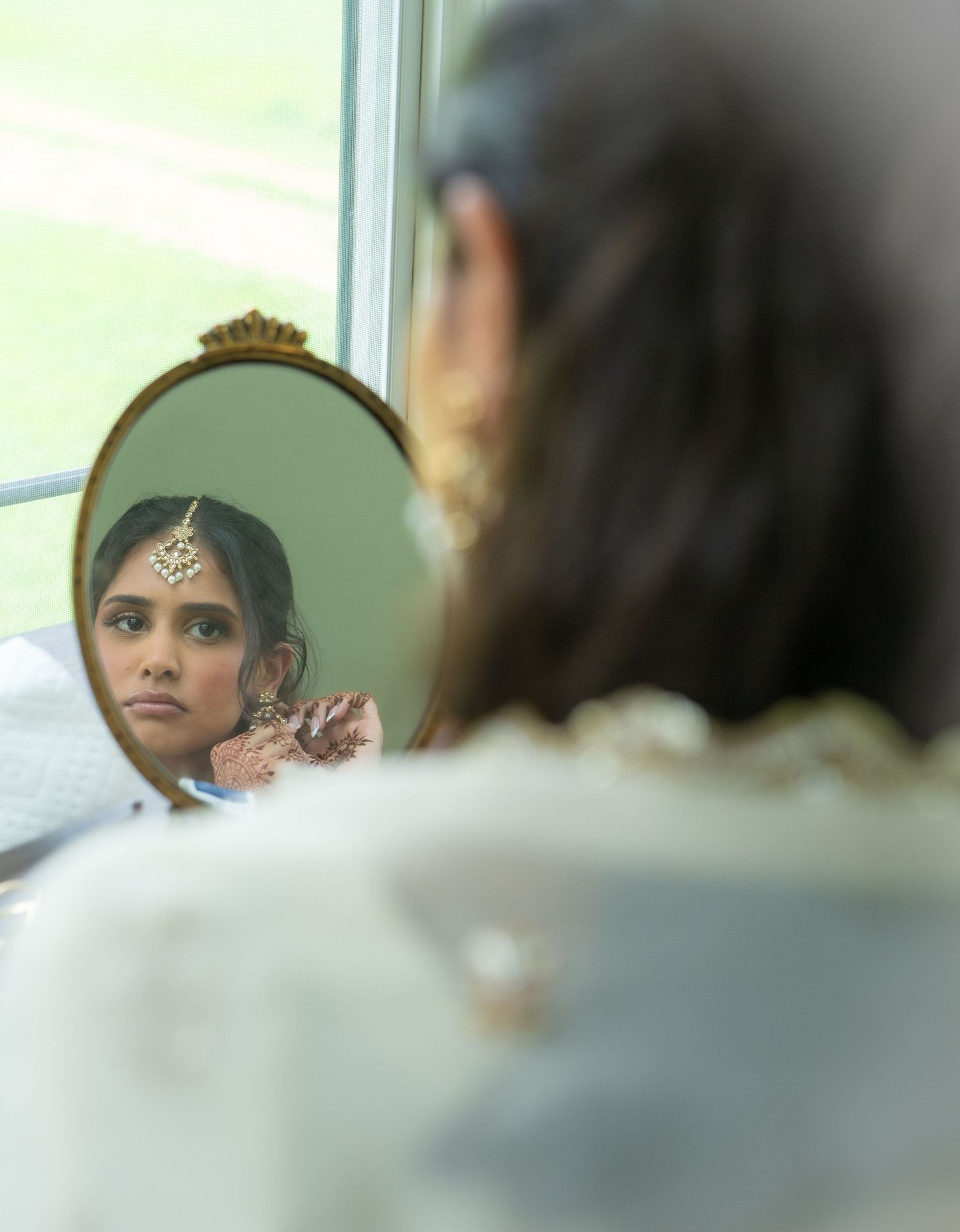 Indian bride adjusting earring while viewing reflection in ornate oval mirror during bridal preparation.
