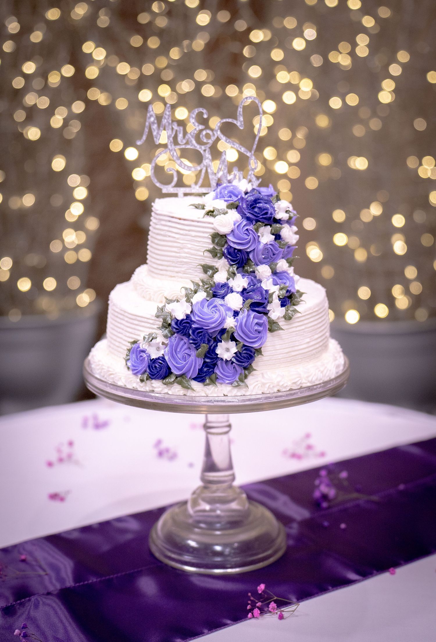 Three-tier white wedding cake with purple roses and Mr & Mrs topper on bokeh background.