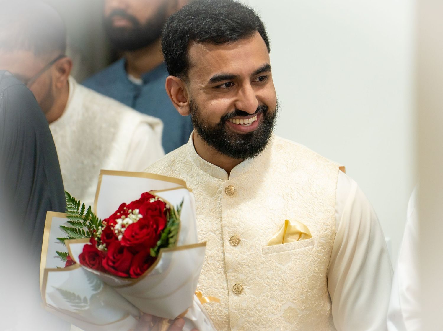 Smiling South Asian man in cream embroidered sherwani holding a bouquet of red roses.