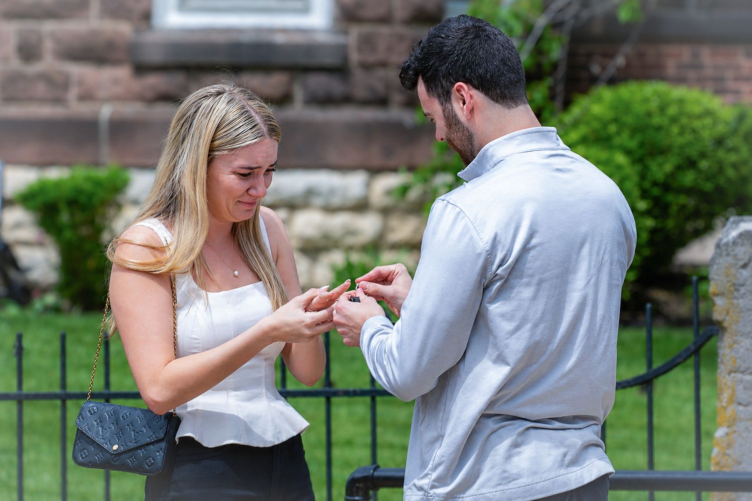 Man placing engagement ring on woman's finger outdoors near historic stone building.