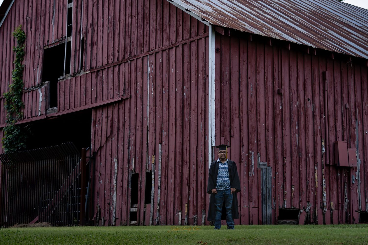 Man standing in front of a large weathered red barn on a green lawn in a rural countryside setting.