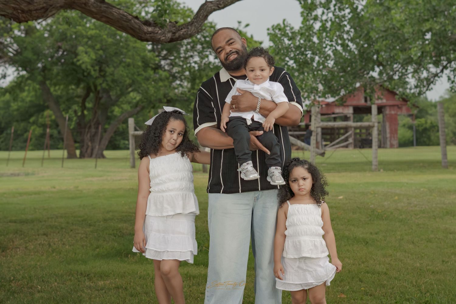 A man holds a baby while posing with two young girls in white dresses on a green lawn near a barn.