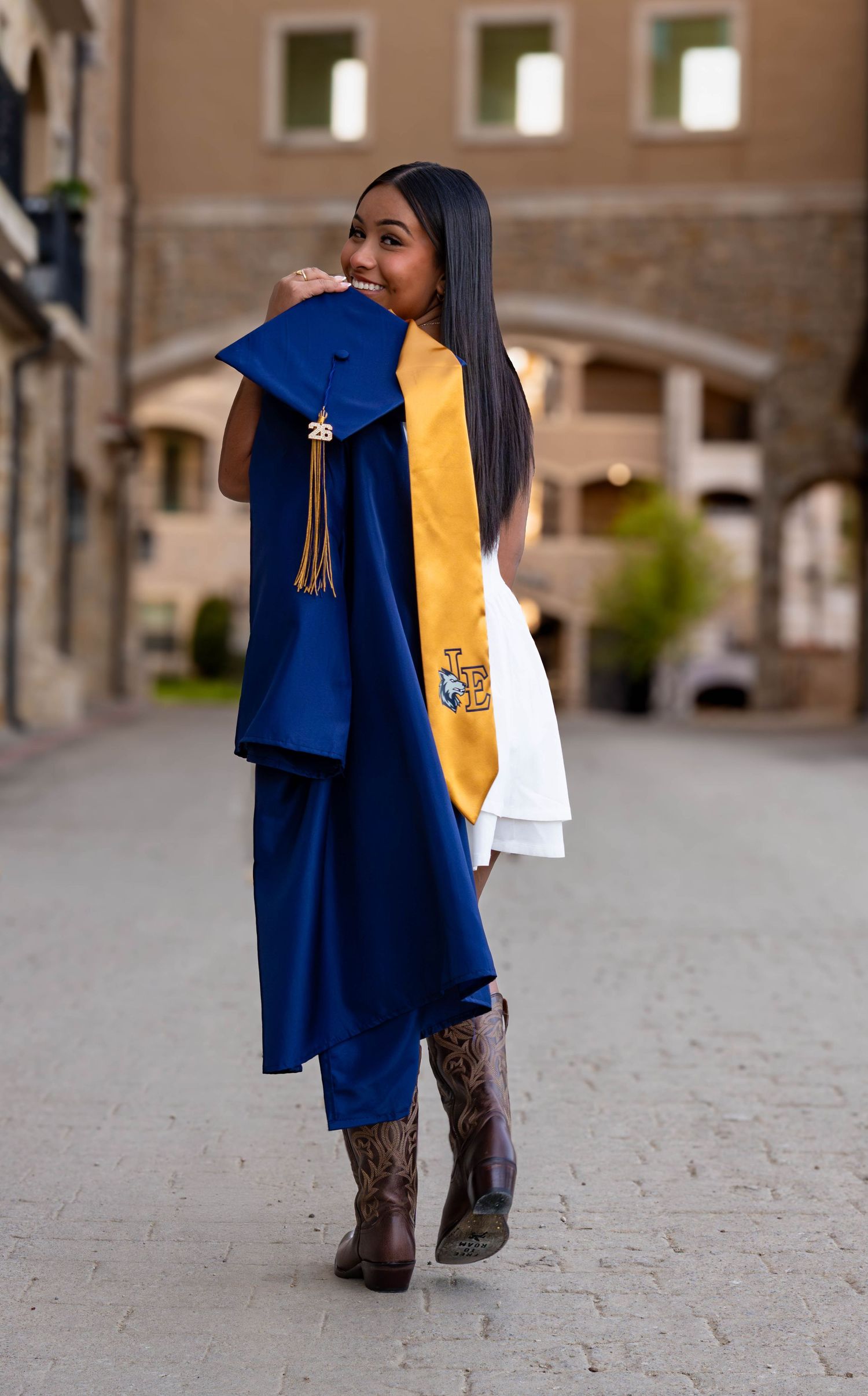 Young woman in blue graduation gown and gold stole holding cap, posing outdoors in cowboy boots.