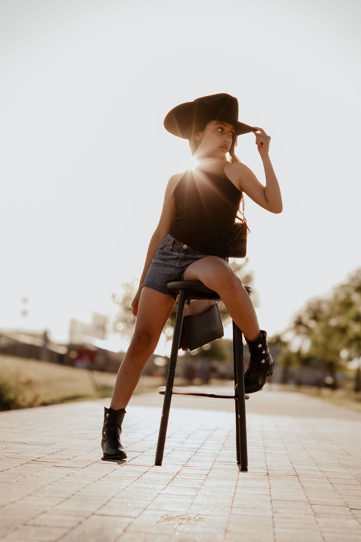 Woman in black hat, top, denim shorts, and boots poses on a stool outdoors in golden sunlight.