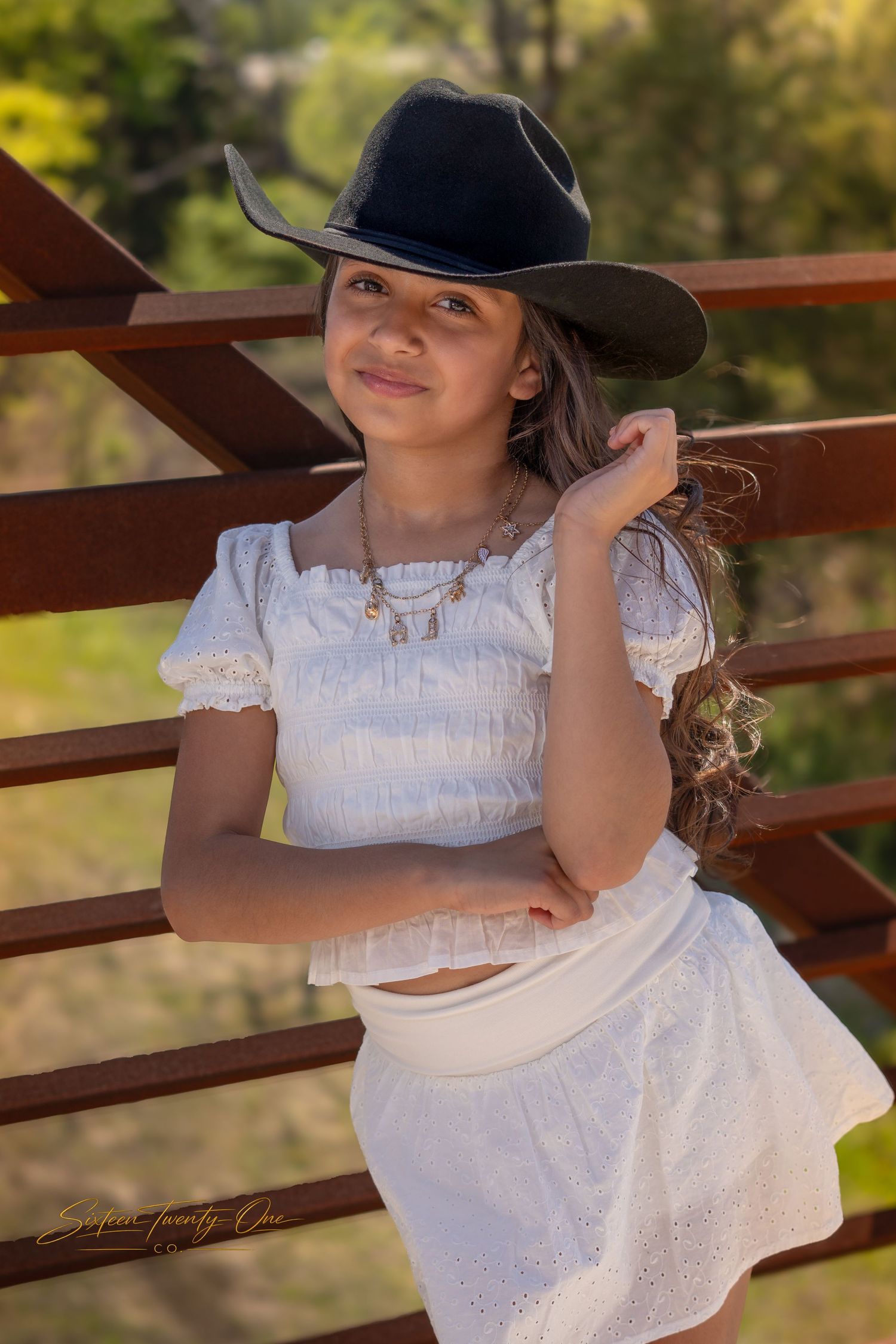 Young woman in white crop top and skirt leans on a rustic fence wearing a black cowboy hat.