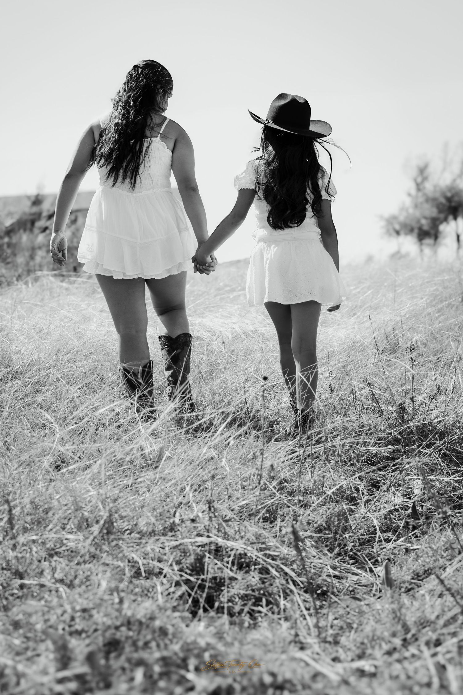 Two young women in white dresses and cowboy boots hold hands walking through a golden grass field in black and white.