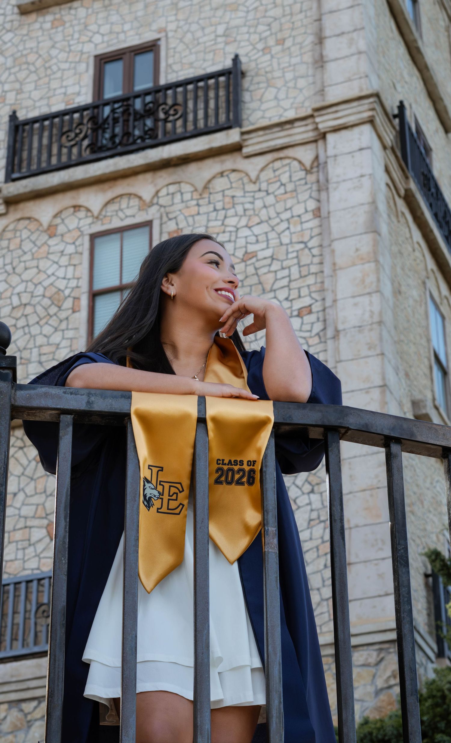 A young woman in navy graduation gown and gold Class of 2026 stole poses by iron fence before stone building.