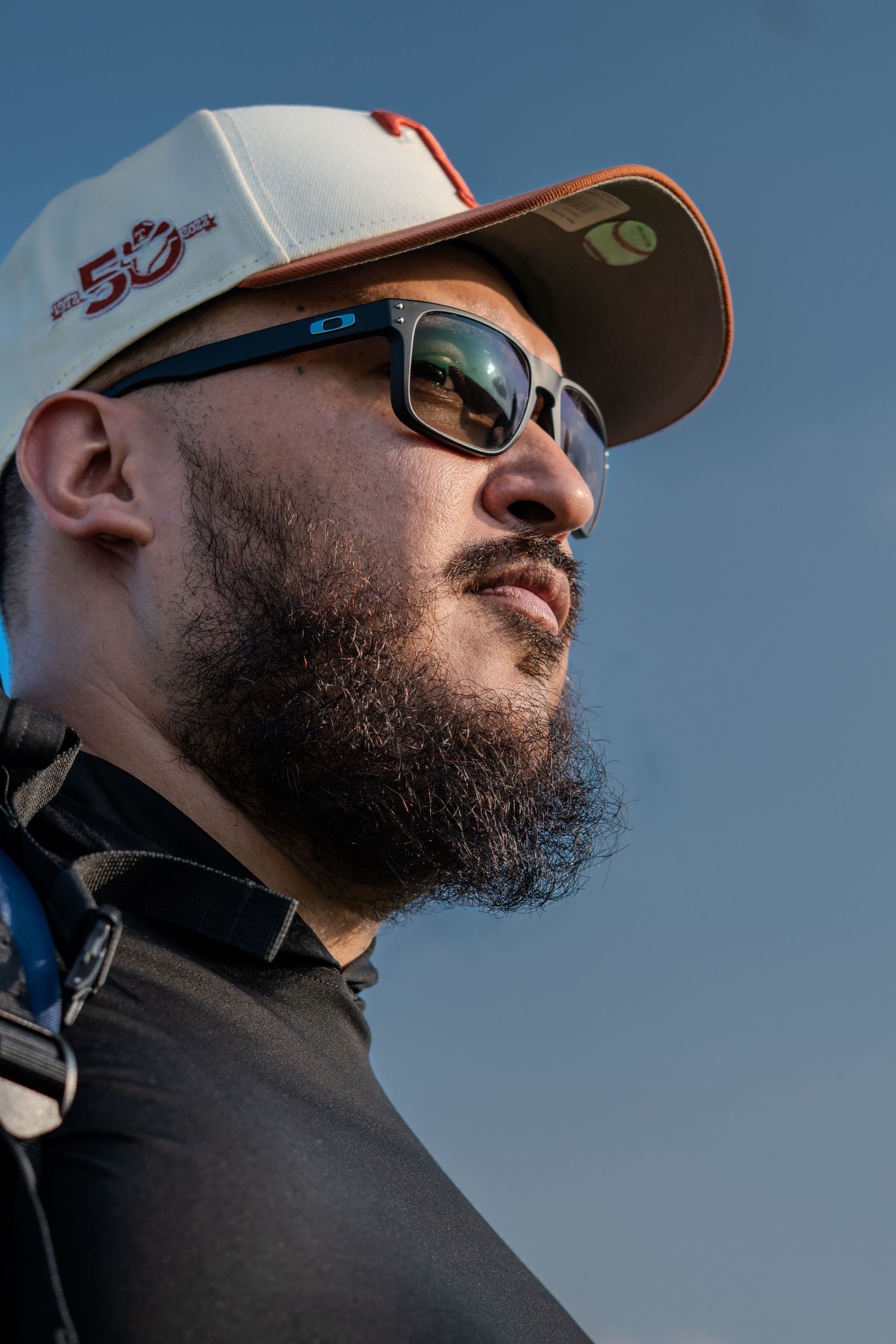 Man with beard wearing Oakley sunglasses and a white baseball cap looks up against a blue sky.