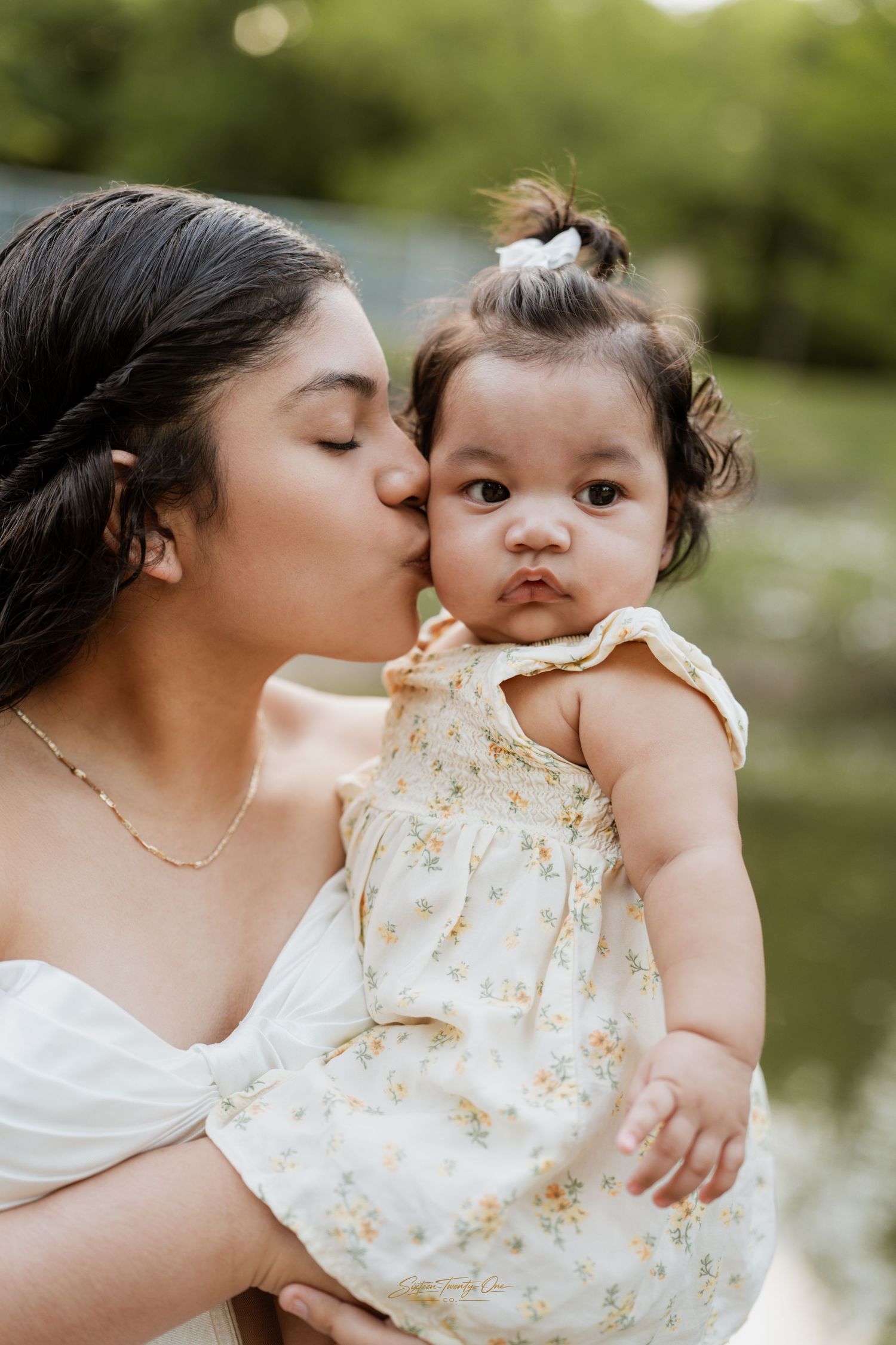 A loving mother kisses her baby girl dressed in a floral romper outdoors near a pond.
