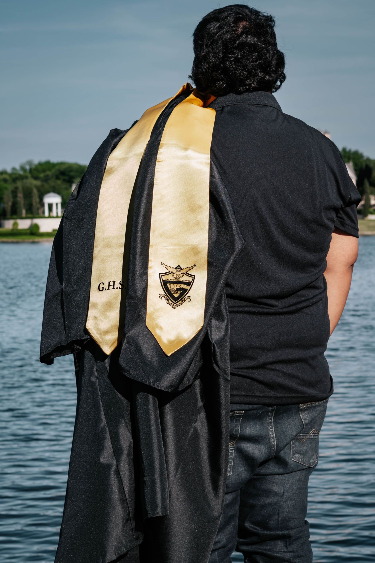 Graduate wearing black cap and gown with gold G.H.S. stole, standing by a lake on a sunny day.