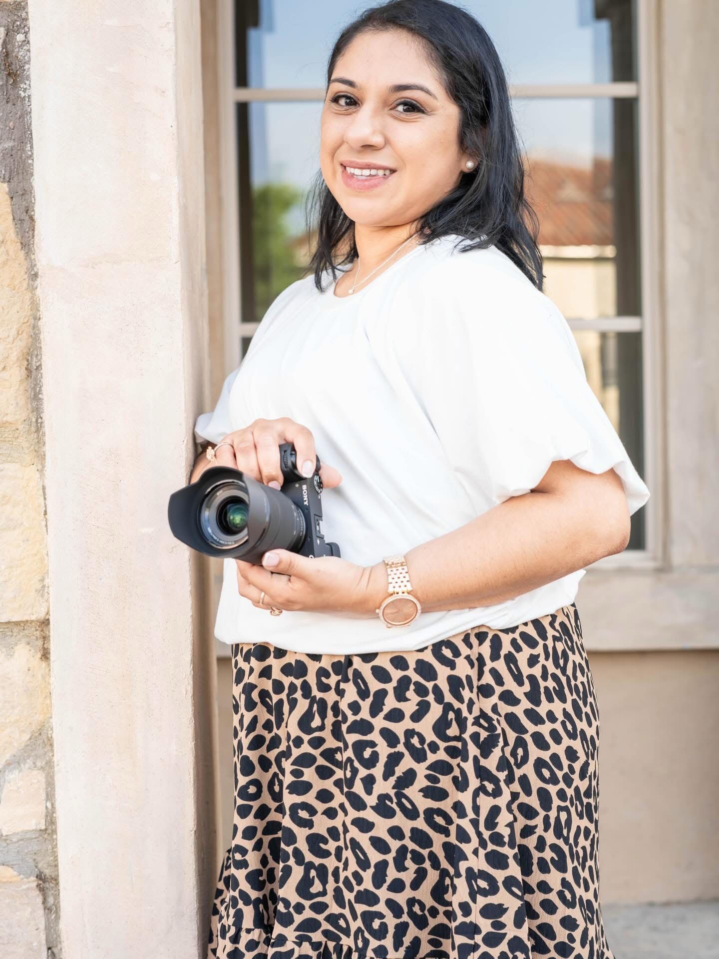 A smiling woman in a white top and leopard print skirt holds a DSLR camera outdoors near a stone building.