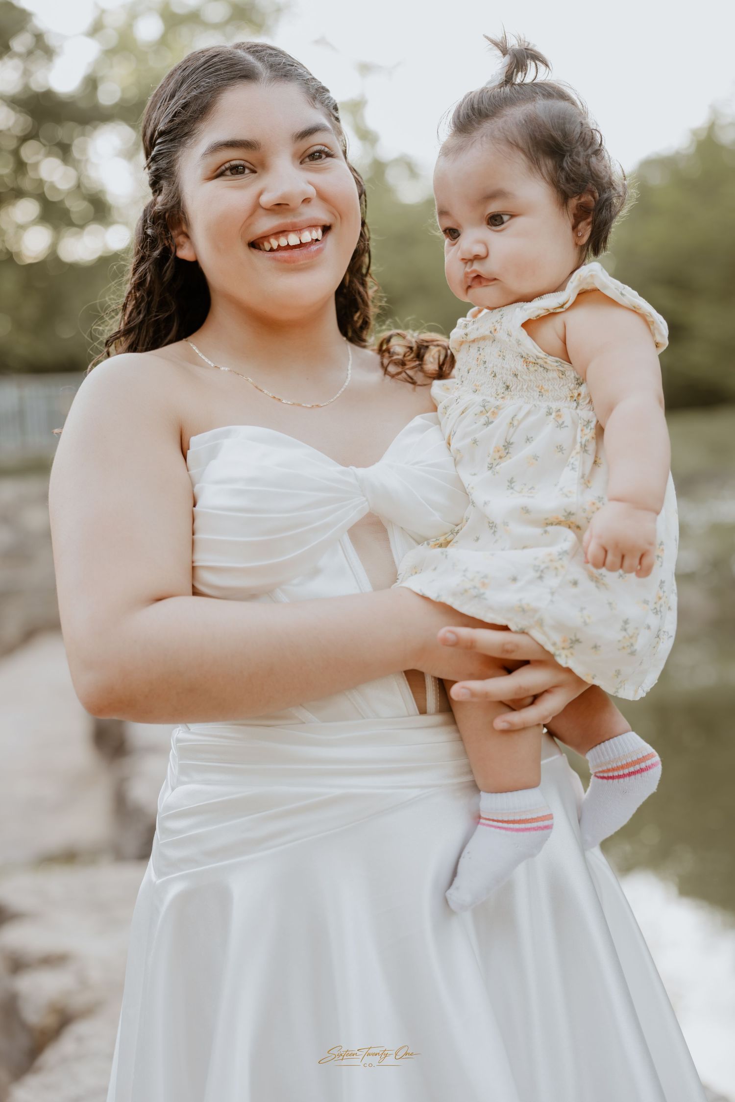 Smiling young woman in white strapless gown holds an adorable baby in a floral dress outdoors.
