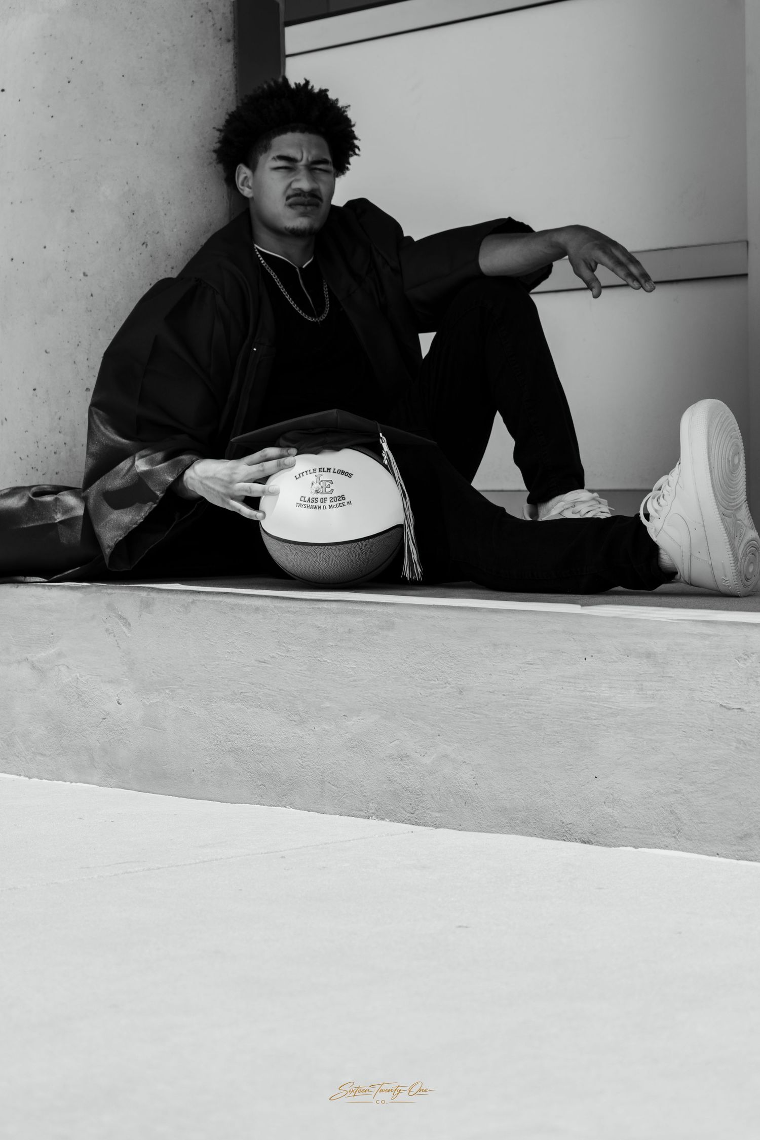 Black and white photo of a young man sitting on steps holding a basketball, wearing all black outfit and white sneakers.