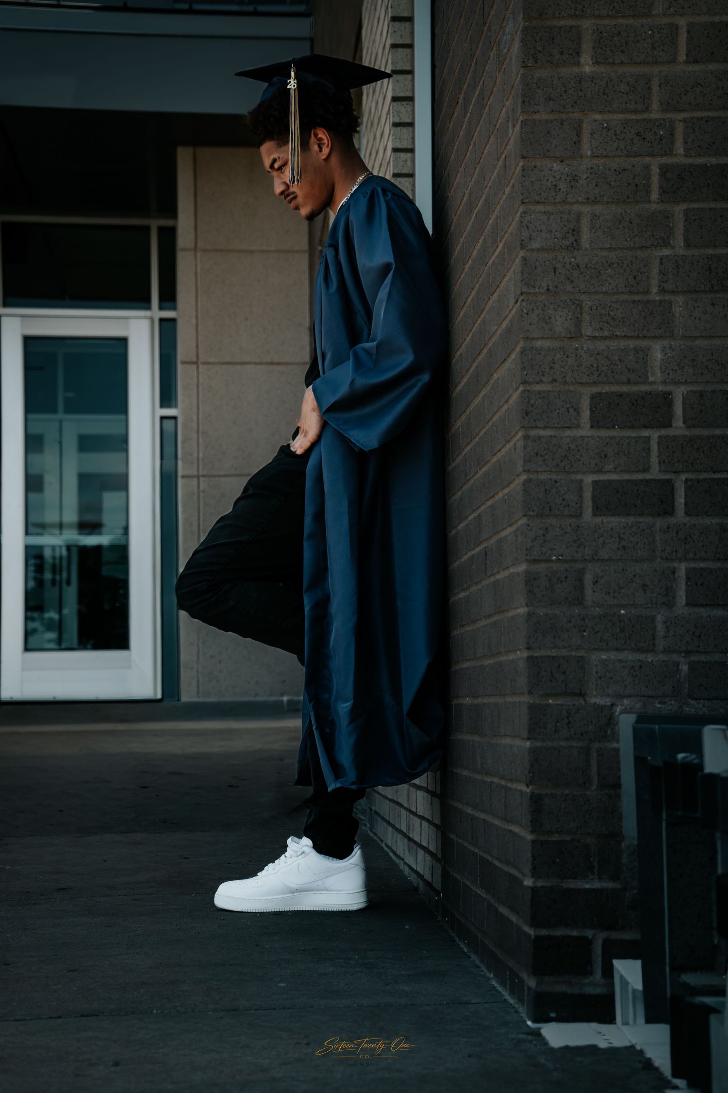 Graduate in blue cap and gown leans against brick wall outdoors, holding mortarboard, wearing white sneakers.