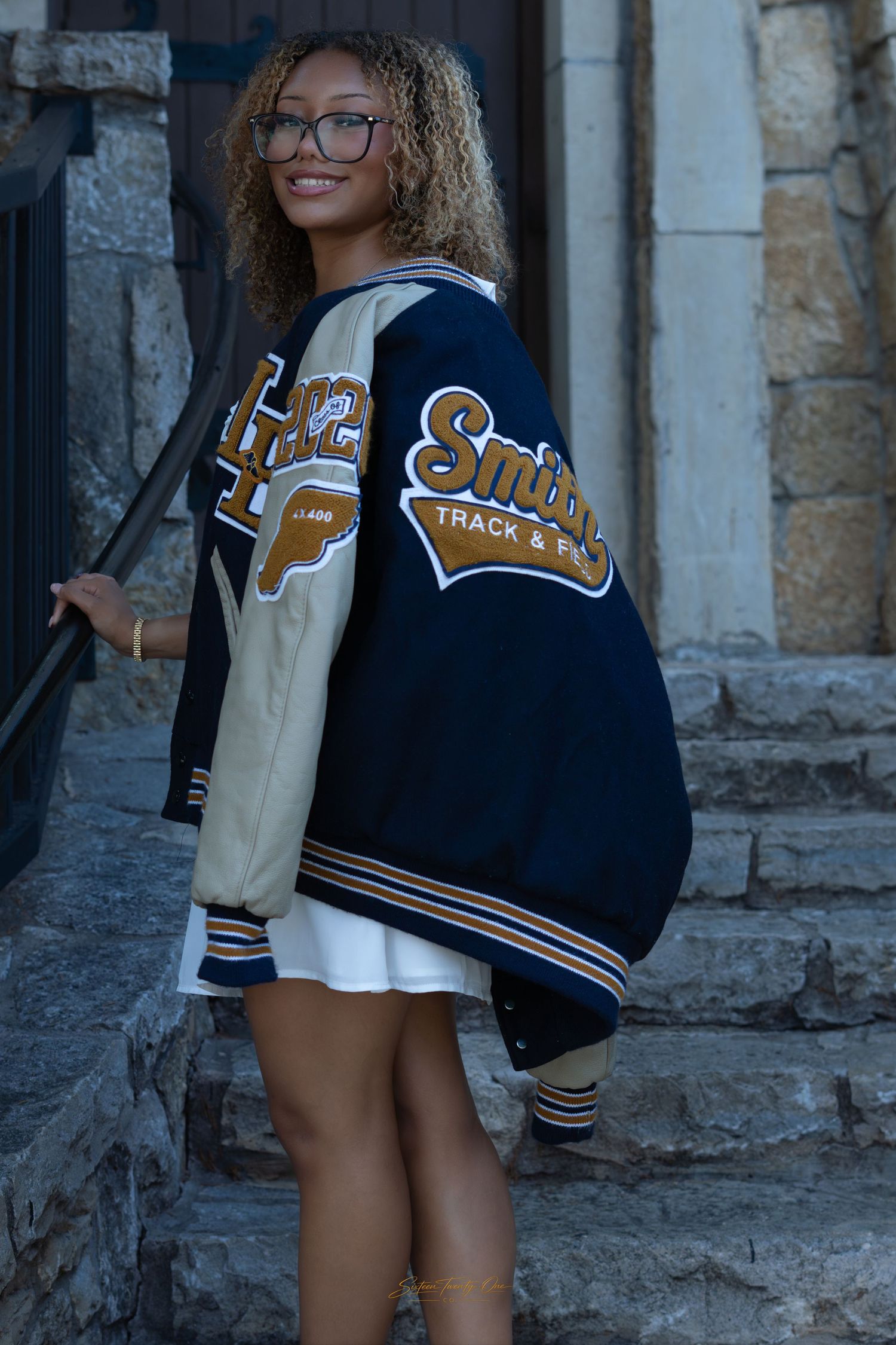Young woman smiling in a navy varsity letterman jacket with Track & Field patches, white dress, on stone steps.
