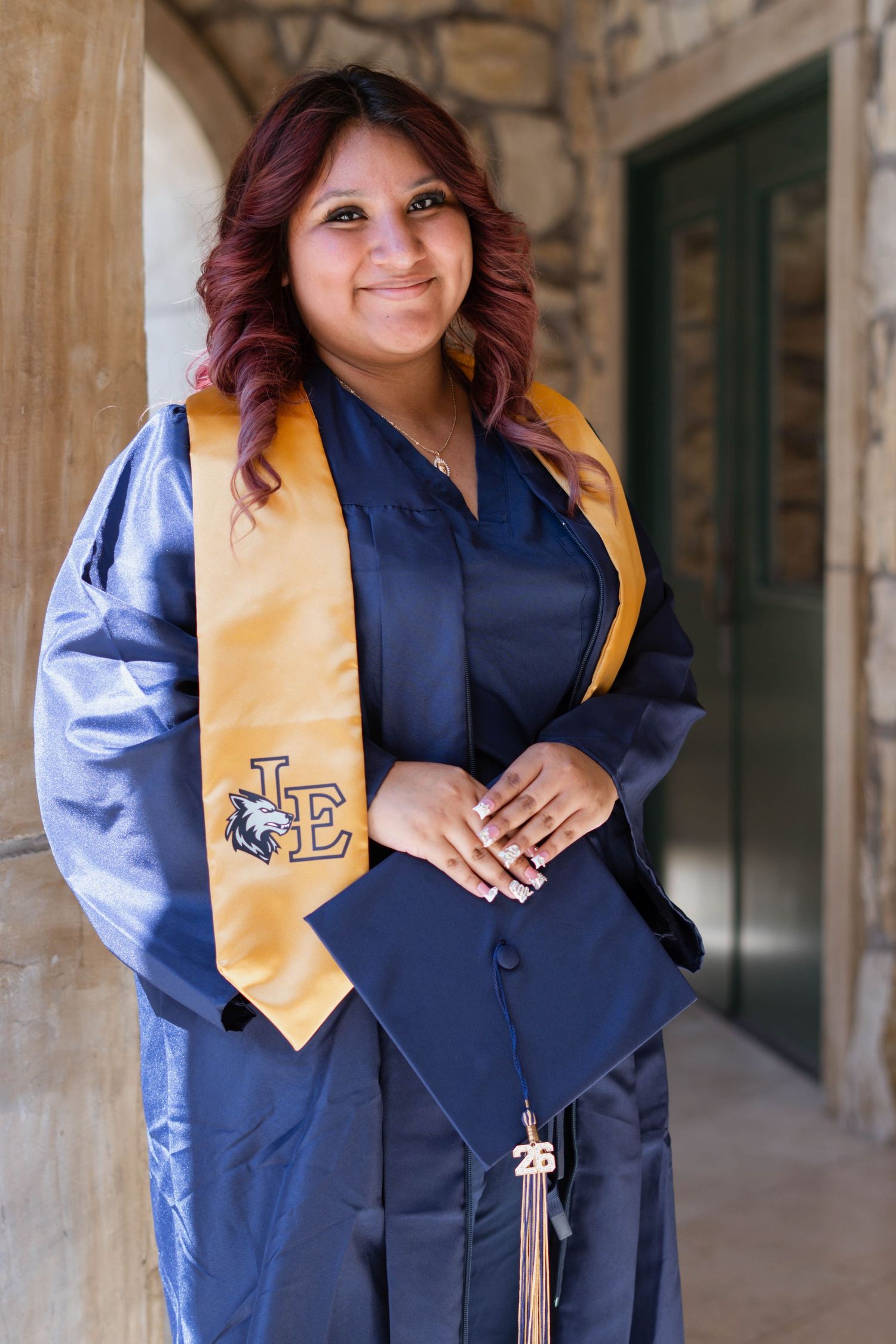 Young woman in navy graduation gown and gold LE stole smiles while posing outdoors for graduation photos.