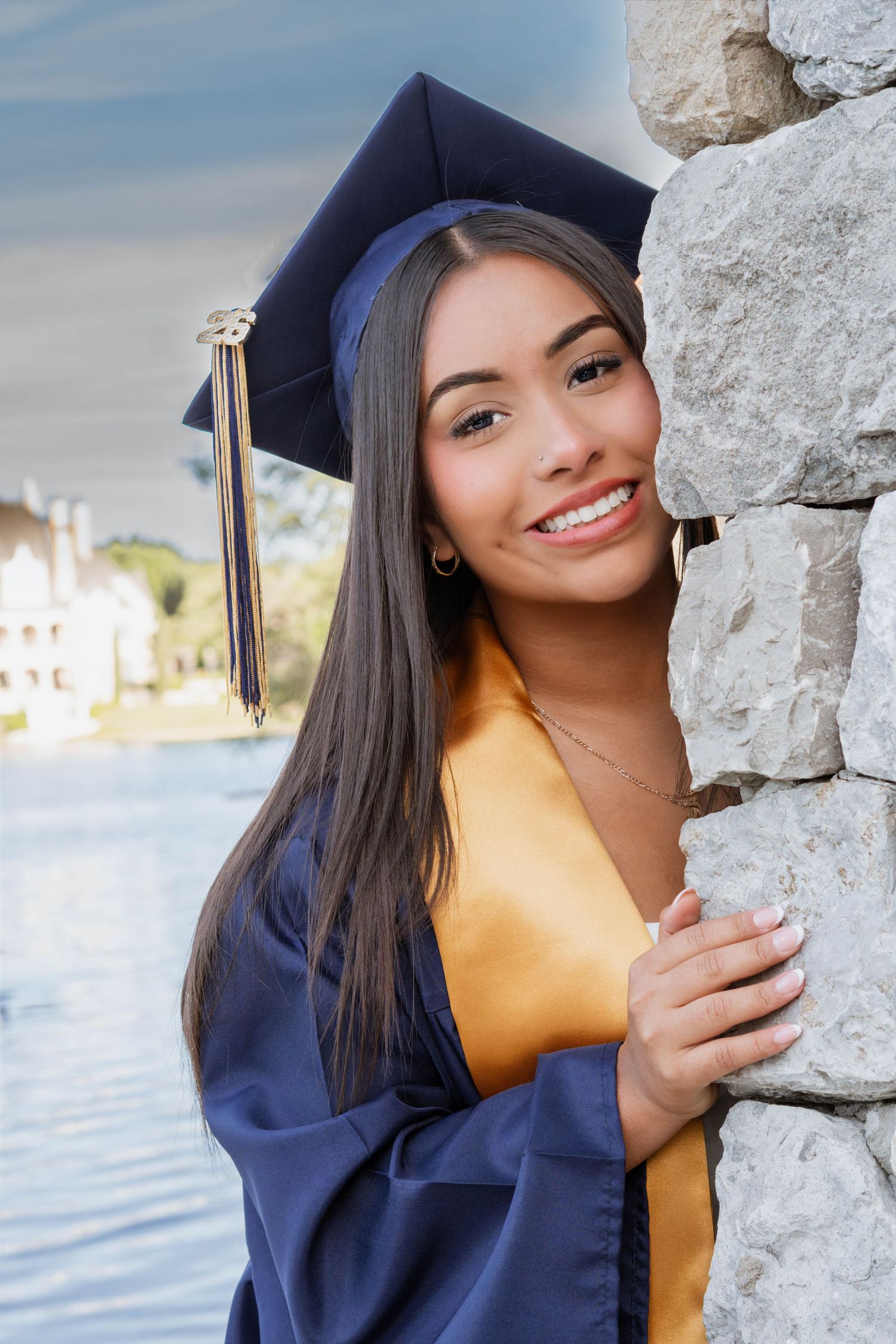 Smiling young woman in navy blue graduation cap and gown posing by a stone wall outdoors.