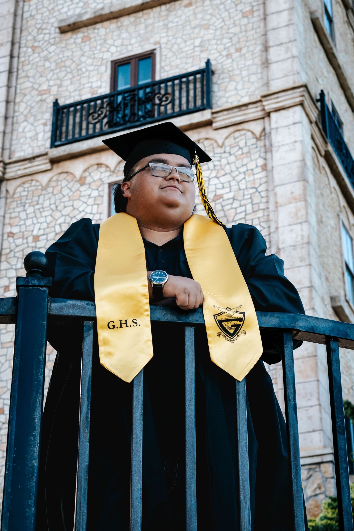 Graduate in black cap and gown with gold GHS stole poses proudly by iron fence in front of stone building.