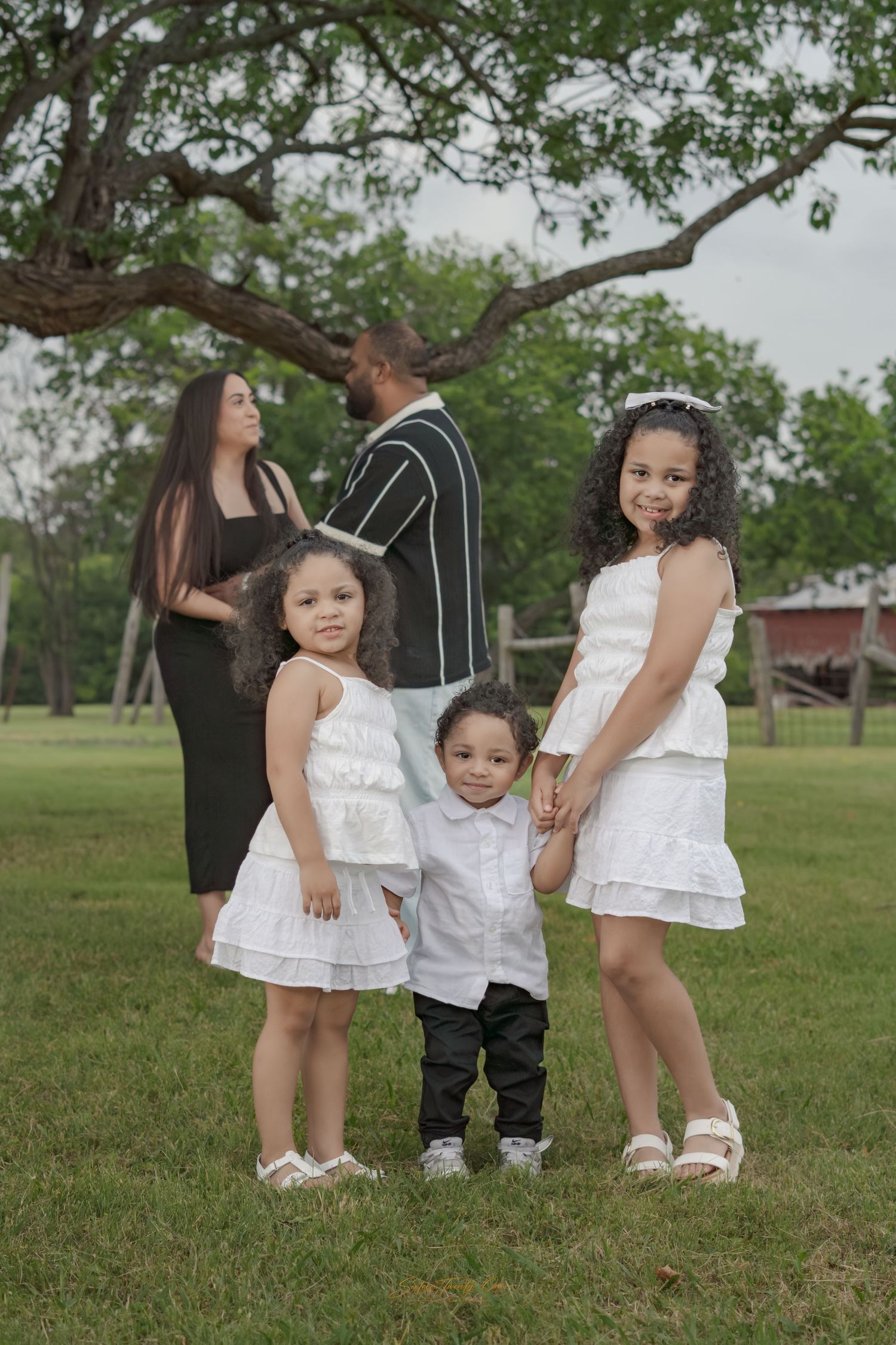 Family portrait with three children in white outfits standing on grass, parents turned away under a large tree.