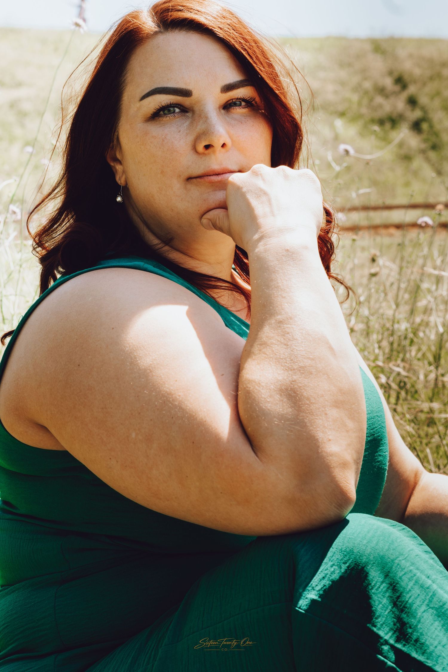 A thoughtful plus-size woman with red hair in a green dress sits outdoors in a sunny field, resting her chin on her hand.