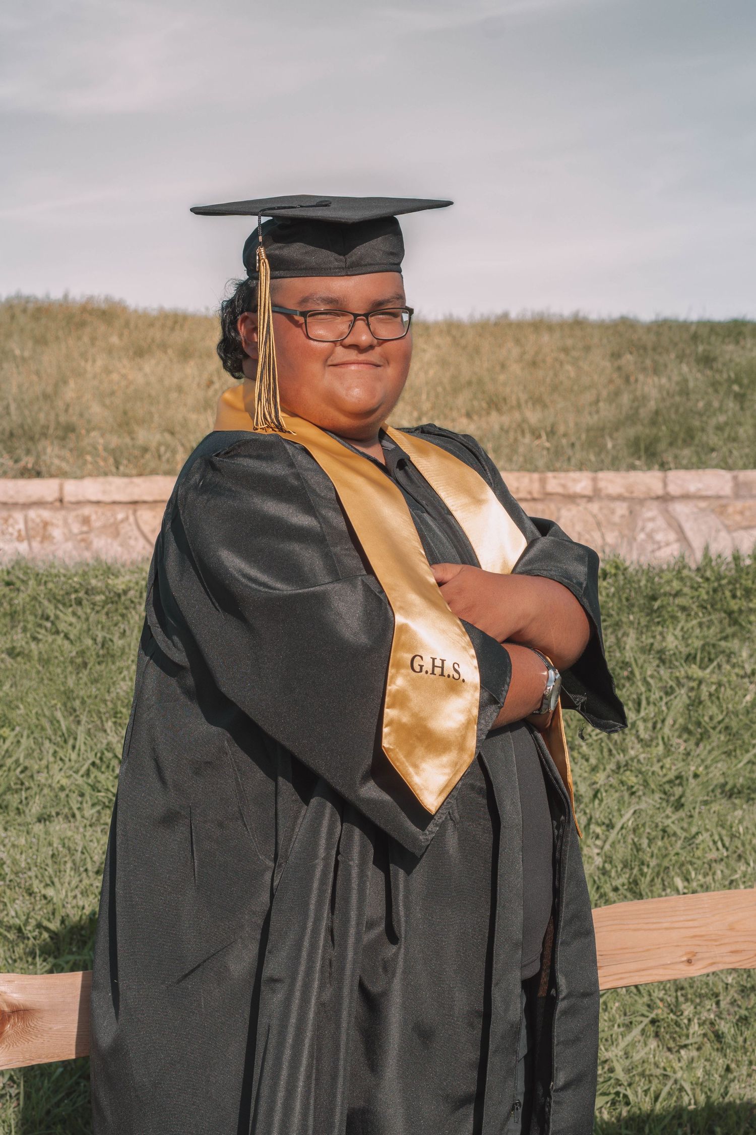 Graduate in black cap and gown with gold GHS stole poses confidently outdoors on a sunny day.