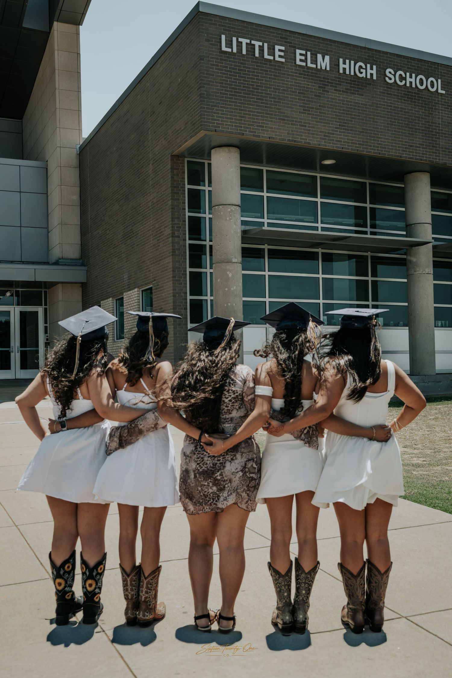 Group of female graduates in white dresses and cowboy boots posing in front of Little Elm High School.