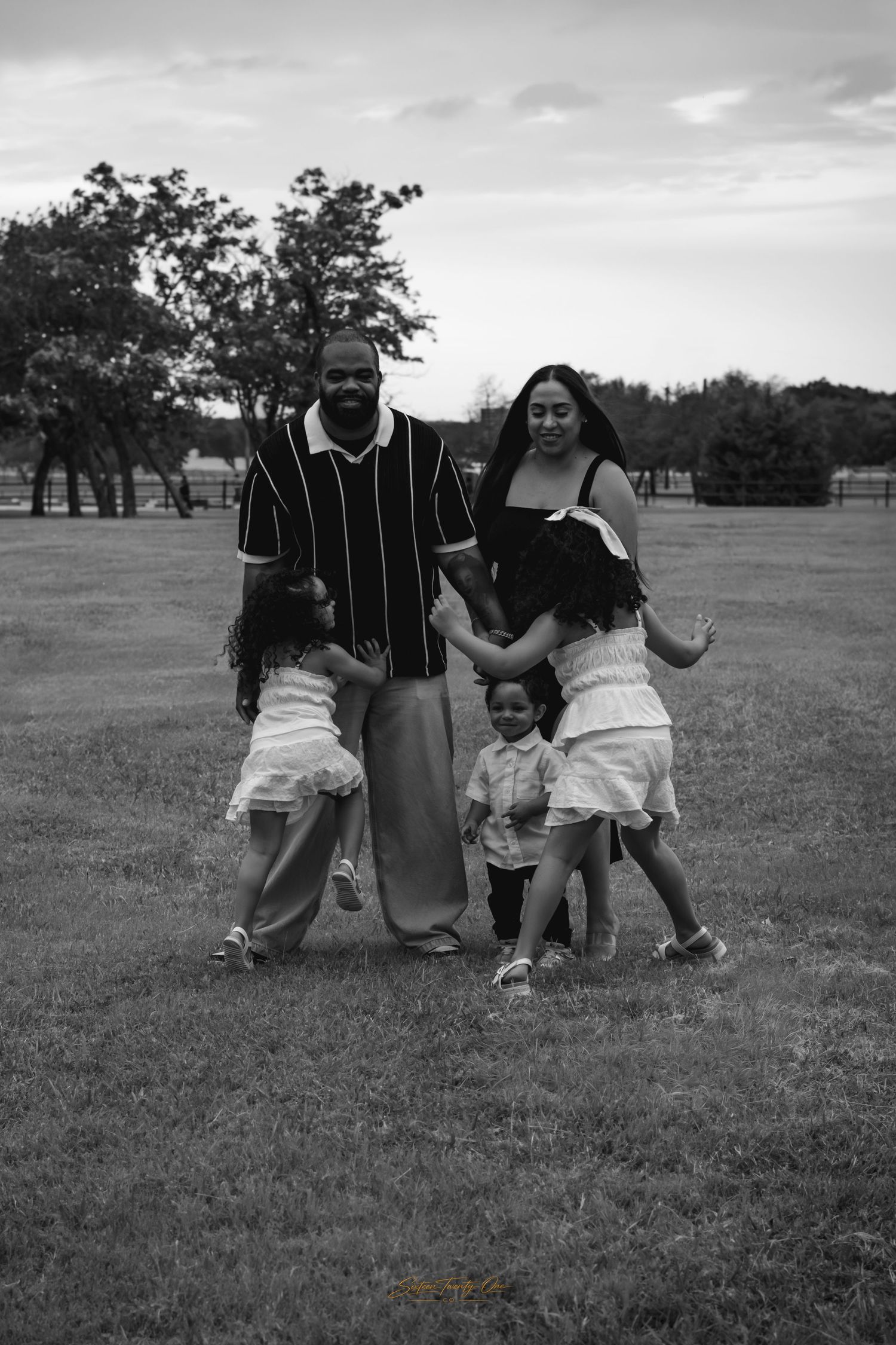 Black and white family portrait of parents with three young children smiling outdoors on a grassy park lawn.