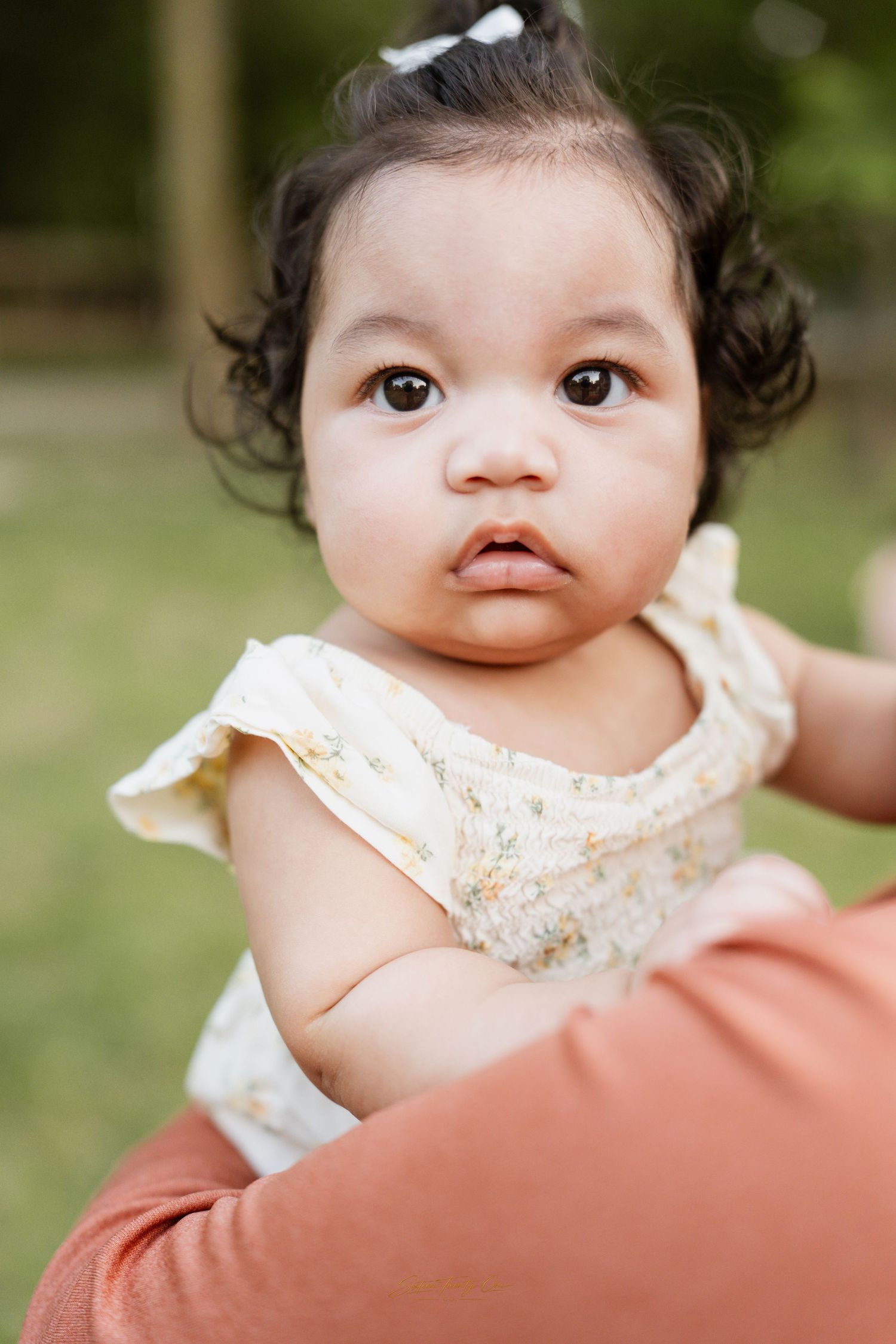 Adorable baby girl in floral dress with dark curly hair and white bow being held outdoors in green park.