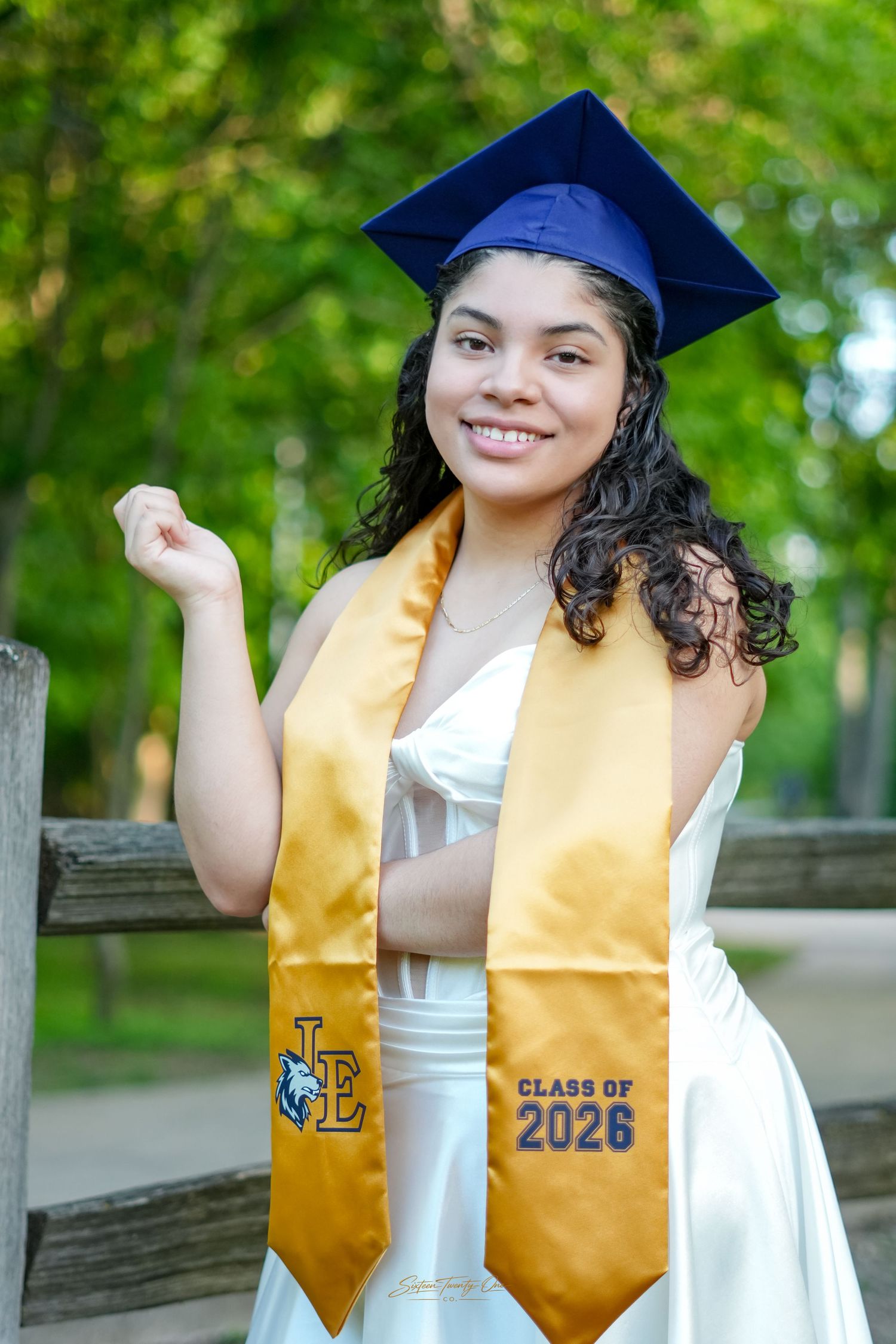 Proud graduate poses in blue cap and gold Class of 2026 stole wearing a white gown outdoors.