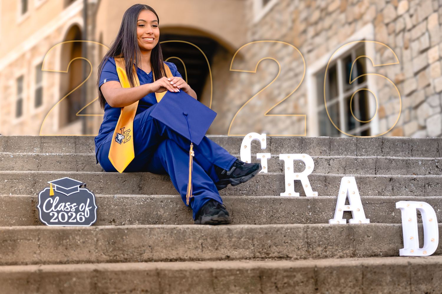 A young woman in blue graduation cap and gown sits on stone steps with GRAD letters and Class of 2026 sign.