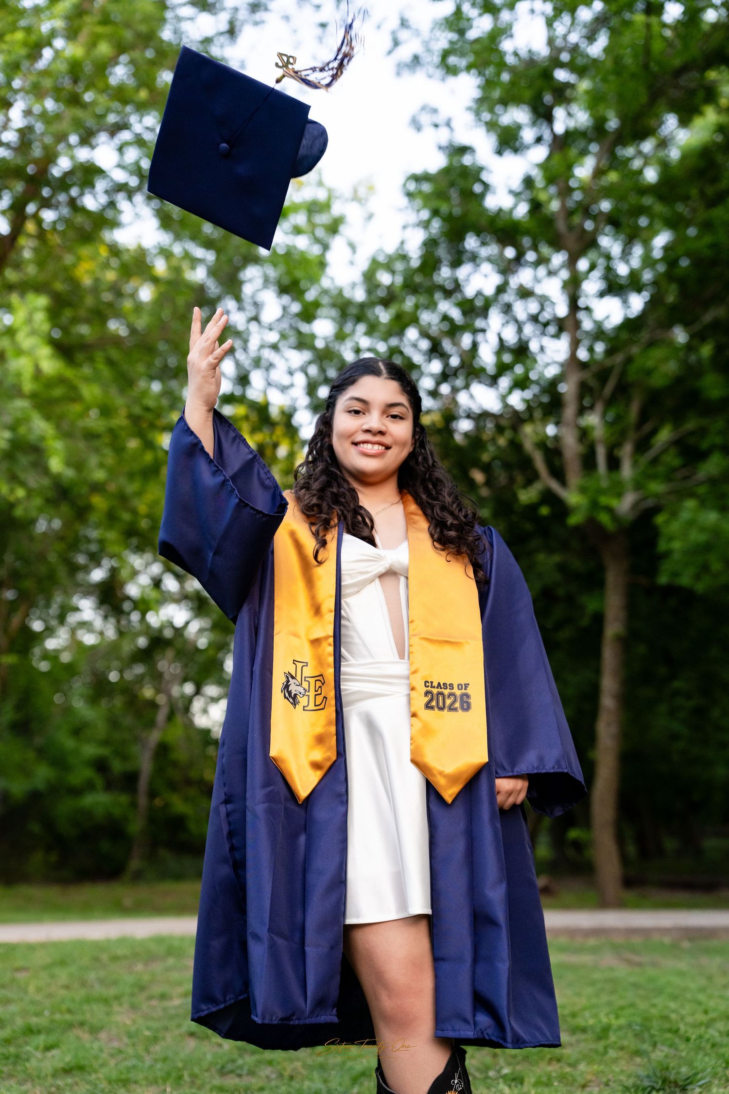 Smiling graduate in navy blue cap and gown tossing mortarboard in air outdoors, wearing Class of 2026 gold stole.