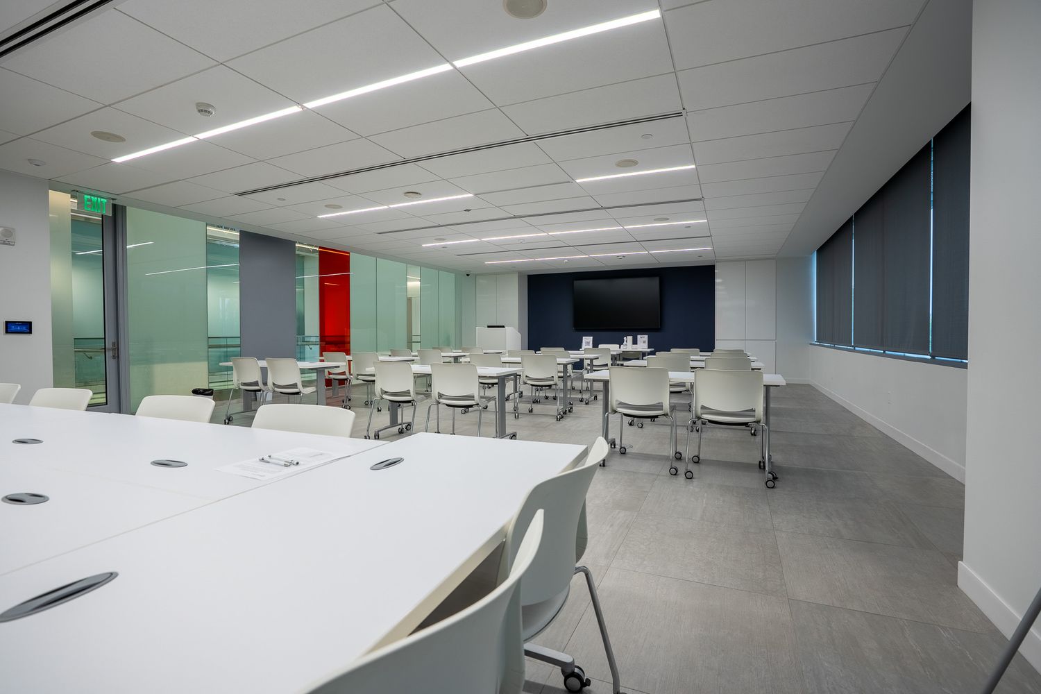 Modern conference room with white tables, chairs, large display screen, and floor-to-ceiling windows in a bright office space.
