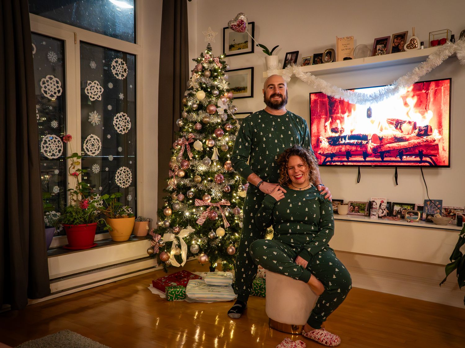 Couple in matching green Christmas pajamas posing by decorated tree in cozy living room with holiday TV display.
