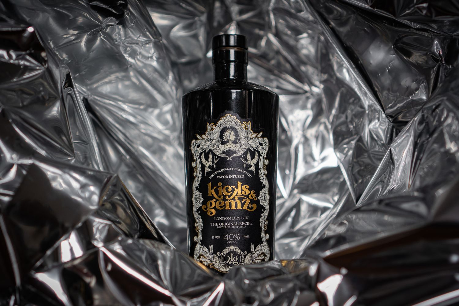 Elegant black bottle of Kicks & Gemz London Dry Gin with ornate gold label on crinkled silver metallic background.