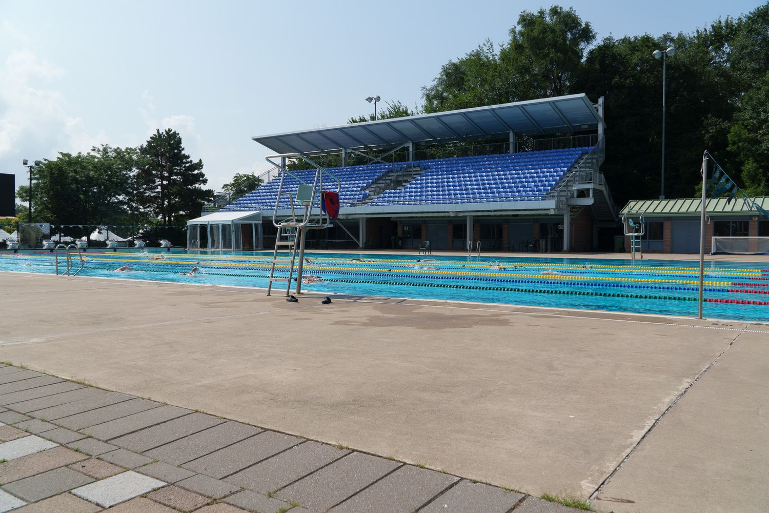Outdoor Olympic swimming pool with blue grandstand seating, lane markers, and lifeguard chair on sunny day.