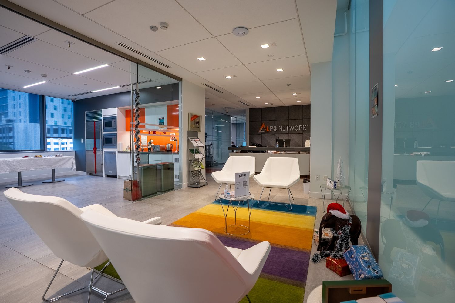 Modern office lounge with colorful rainbow rug, white chairs, glass walls, and vibrant open workspace in background.
