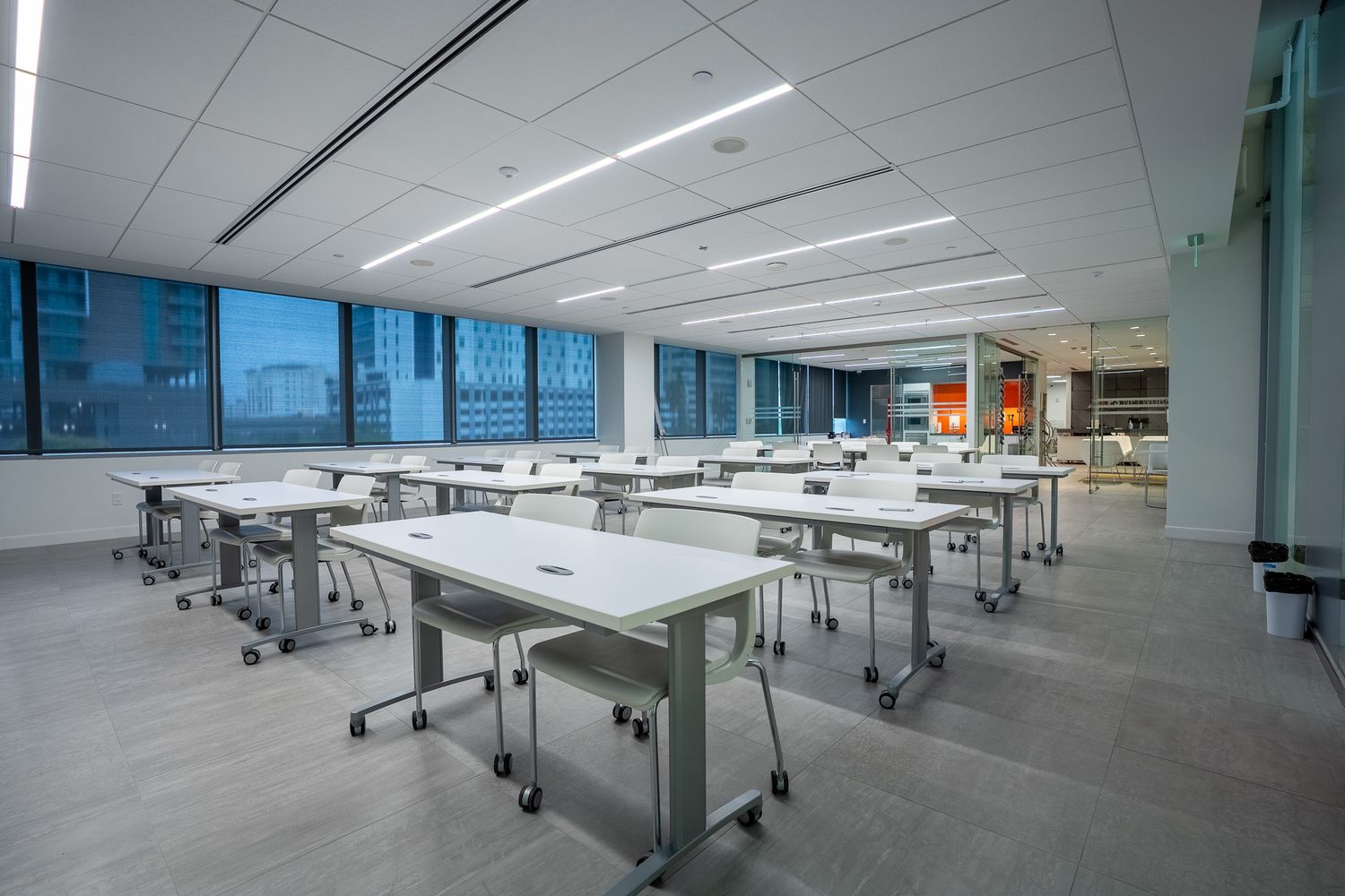 Modern empty classroom with rows of white mobile tables and chairs, large windows, and bright LED lighting.