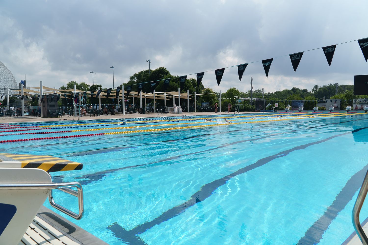 Outdoor Olympic swimming pool with blue lane lines, black triangle flags, and overcast skies at an aquatic sports facility.