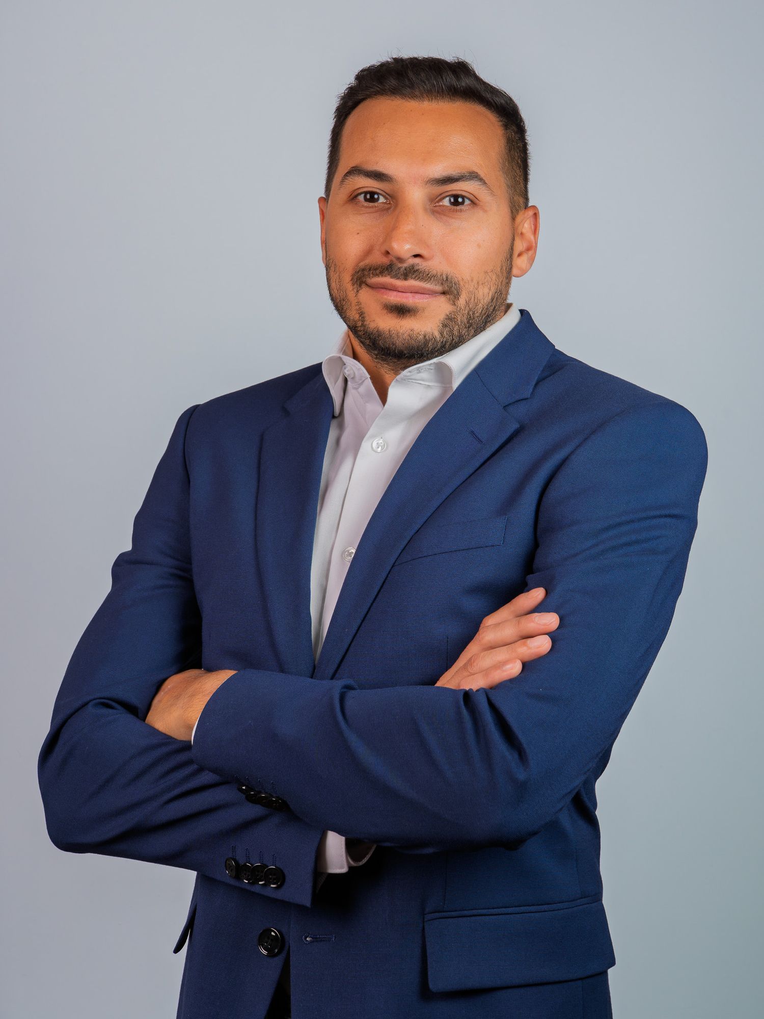 Professional man in navy blue suit with arms crossed poses for a corporate headshot on gray background.